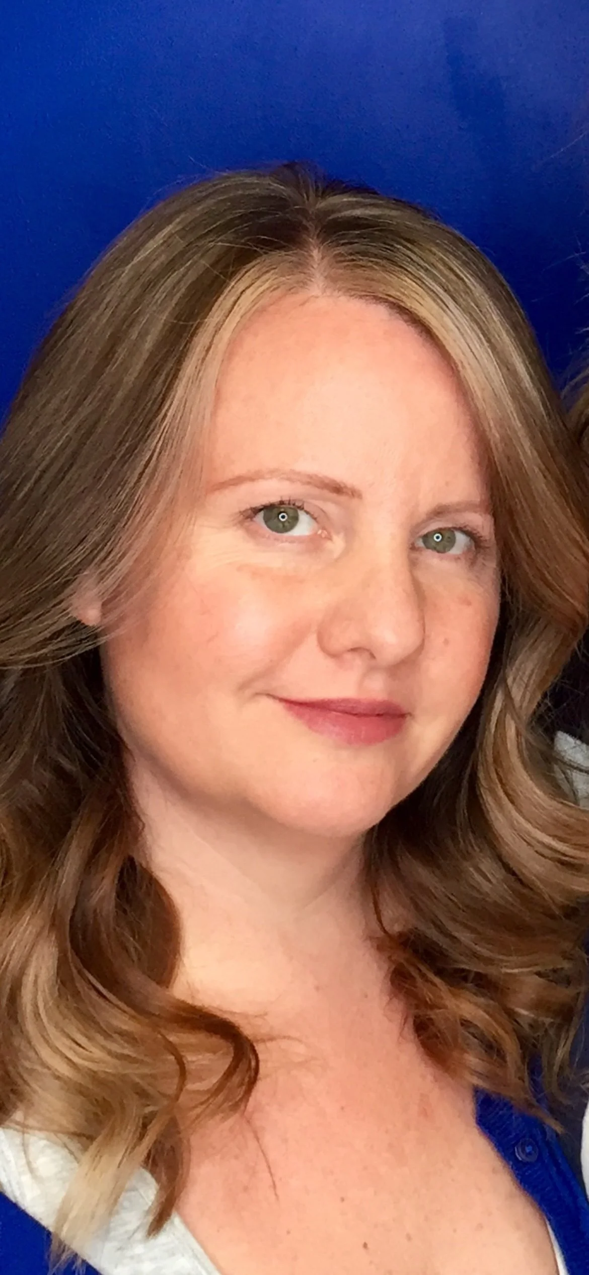 Close-up of Kim smiling with wavy light brown hair, wearing a blue and white shirt, against a blue background.