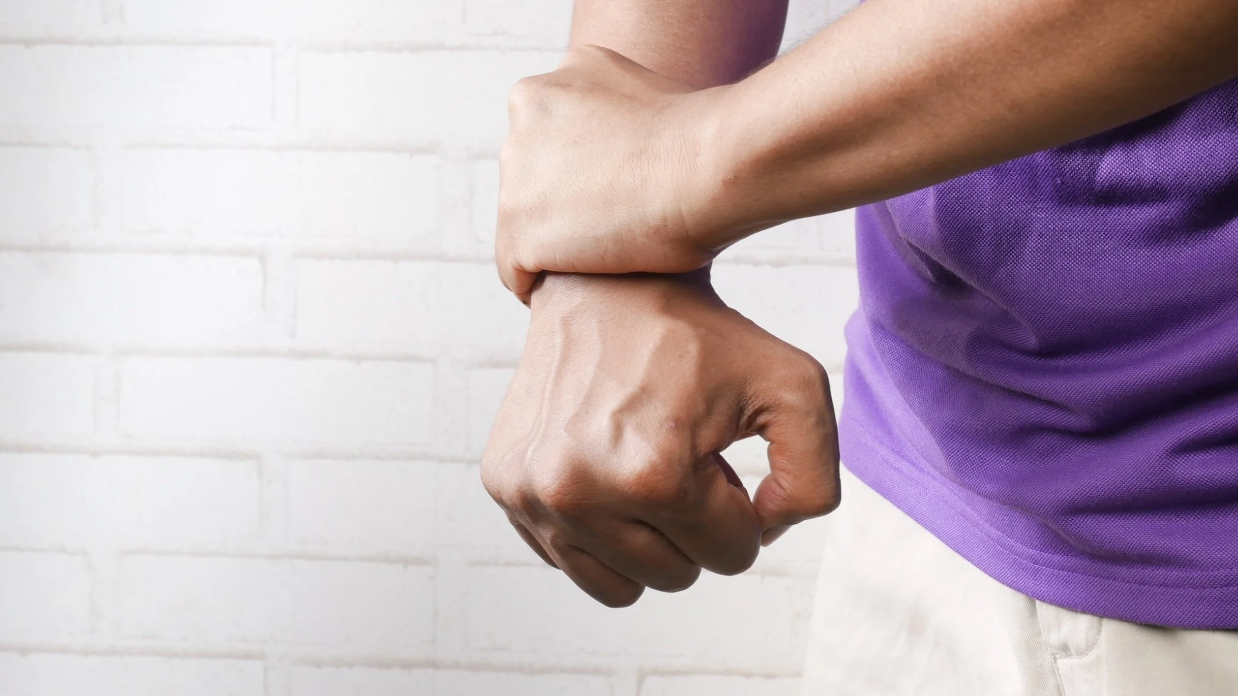 Person holding or embracing with their hand wrapped around their wrist against a white brick wall background.