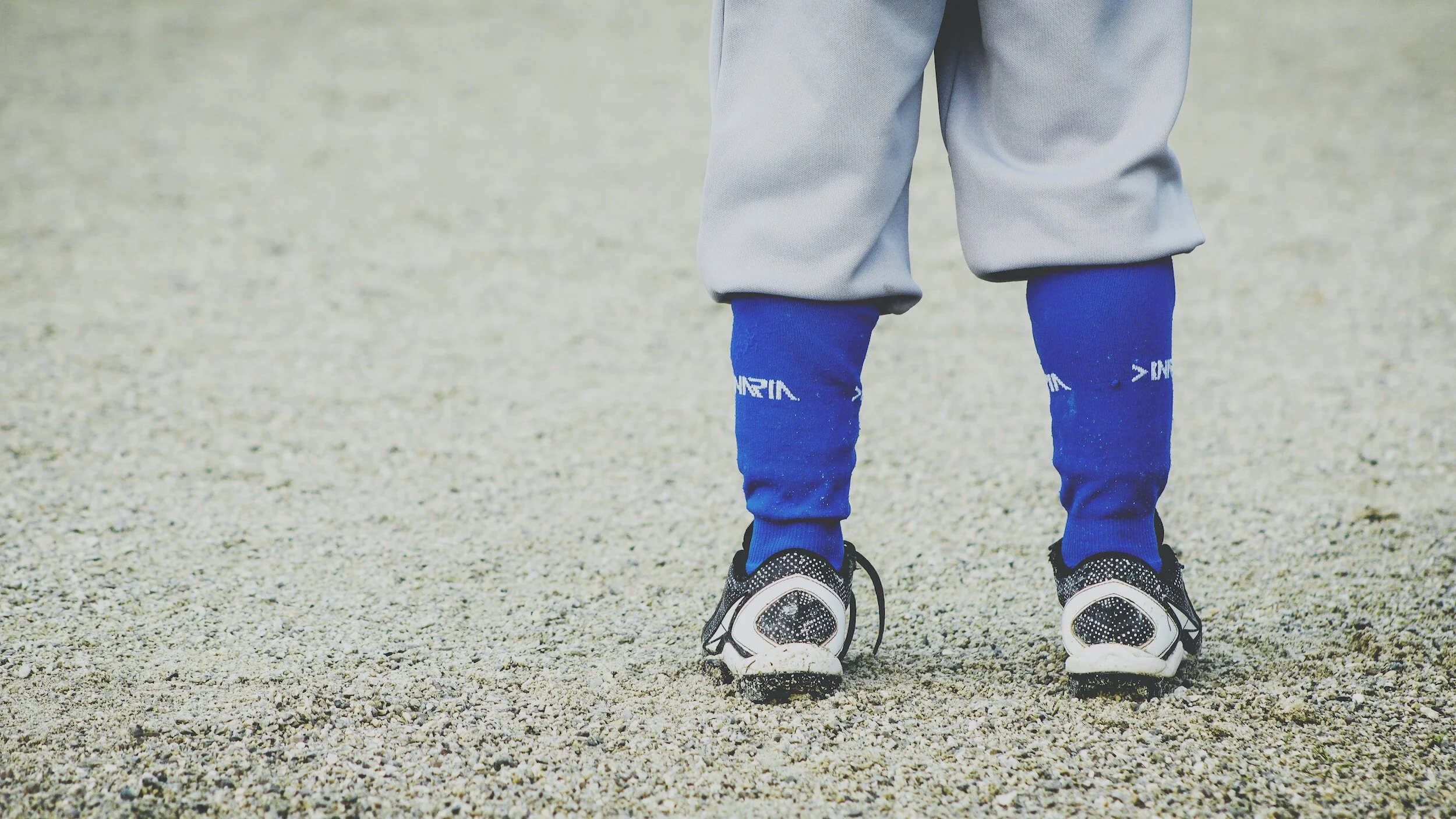 Child wearing black and white athletic shoes, blue socks, gray pants, and standing on gravel surface.