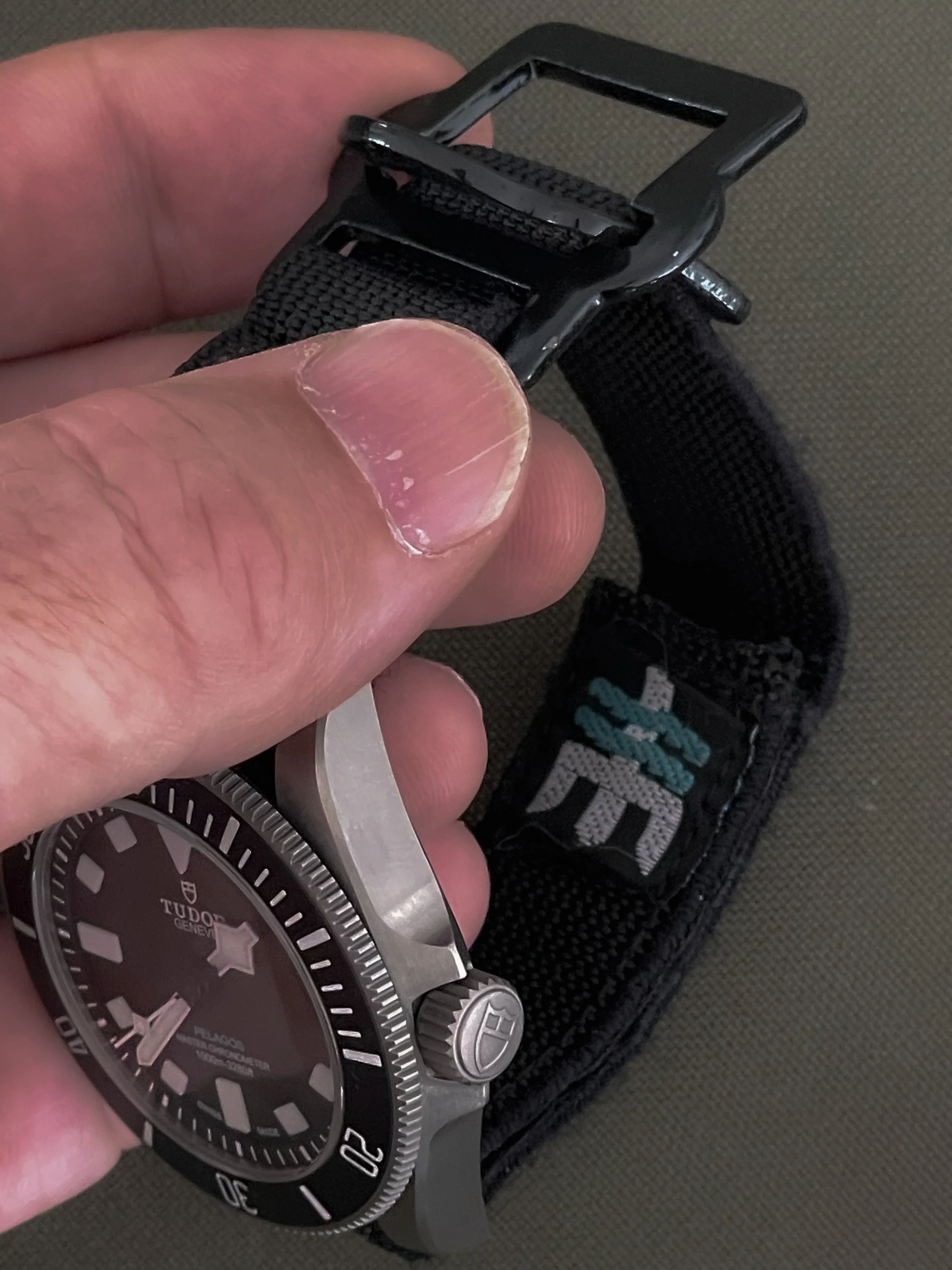 A hand holding a wristwatch with a black strap and a metal case, attached to a black strap with a black buckle. The background is a textured surface.