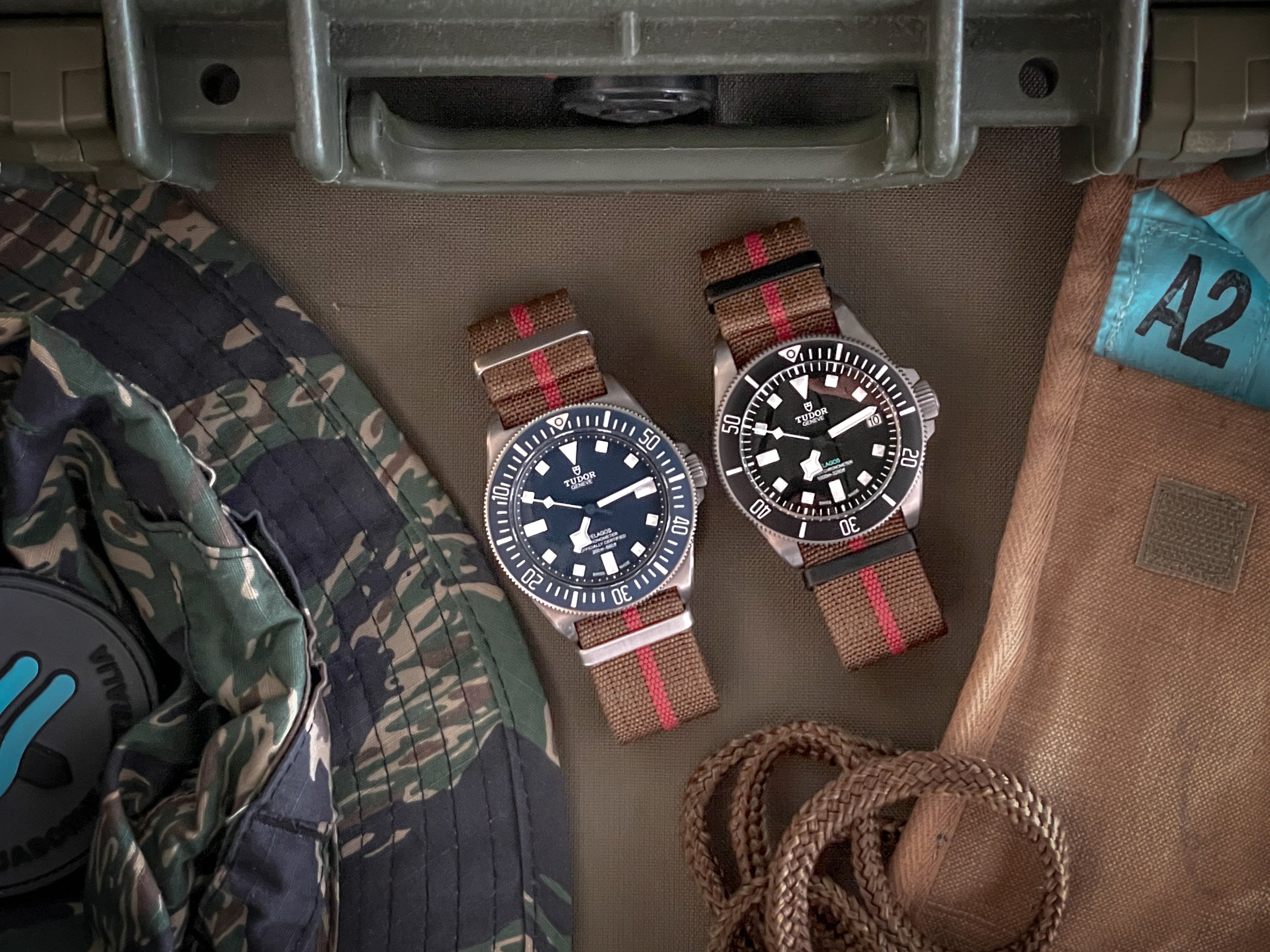 Two Tudor dive watches with black dials and brown fabric straps, positioned on a beige surface, surrounded by camouflage clothing, a brown pouch, and a pair of brown rope laces.