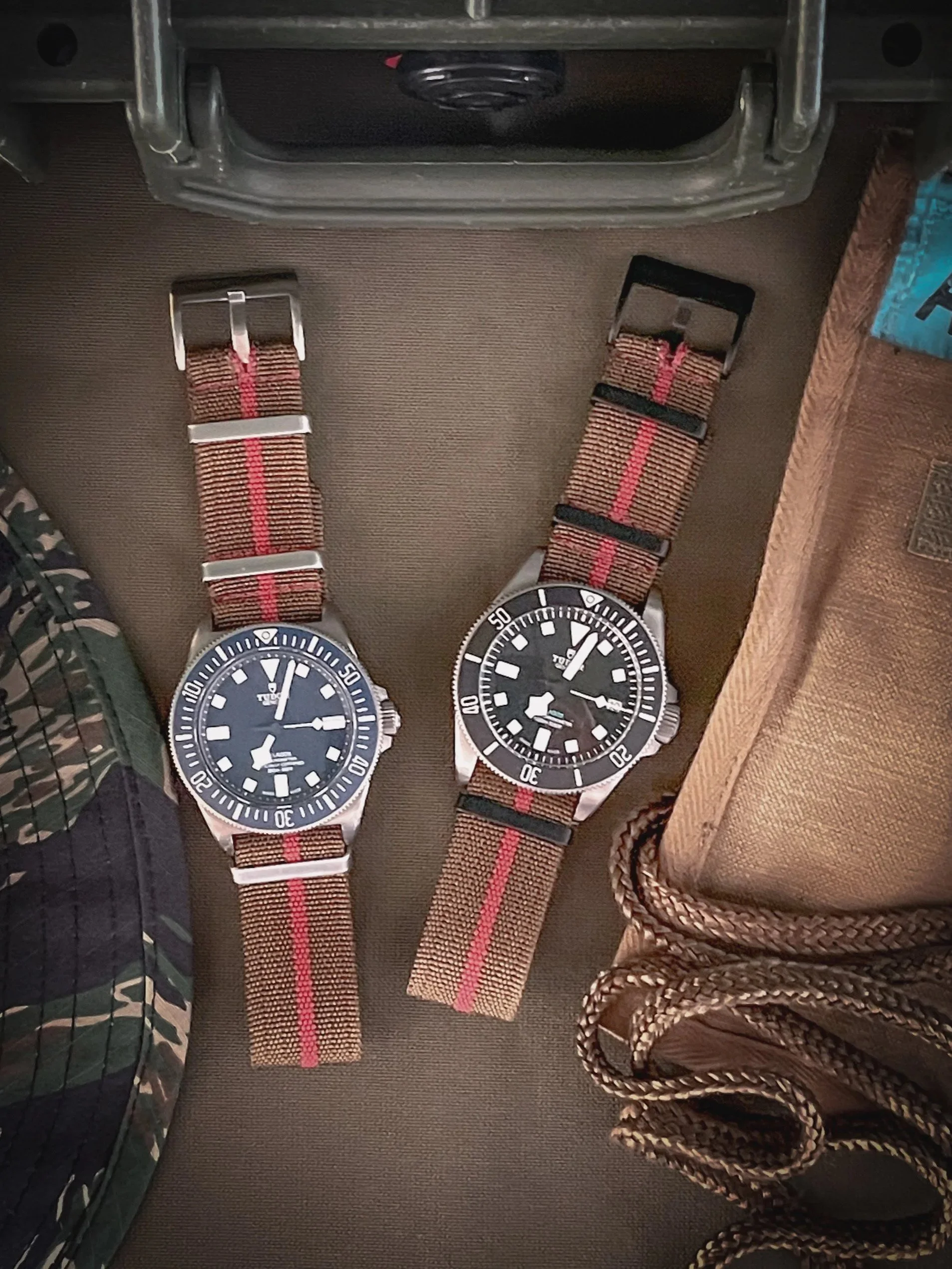 Two wristwatches with brown and red striped NATO straps placed side by side, on a brown surface. The watches have black dials and silver cases, with the left watch showing a 12-hour clock and the right a 24-hour clock and a rotating bezel.