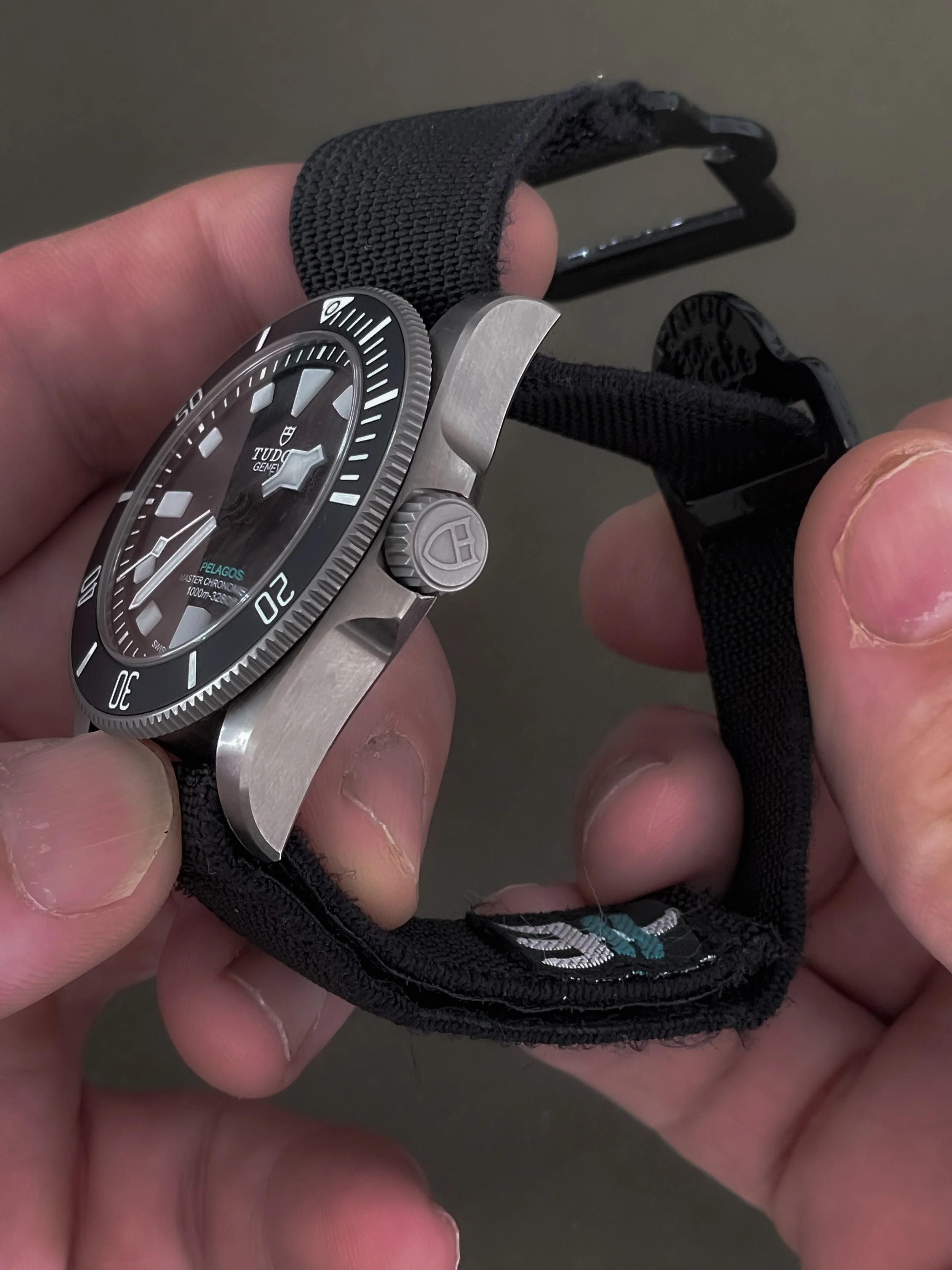 Close-up of a Tudor Pelagos dive watch with a black dial and bezel, worn on a black fabric strap, held in a person's hand.