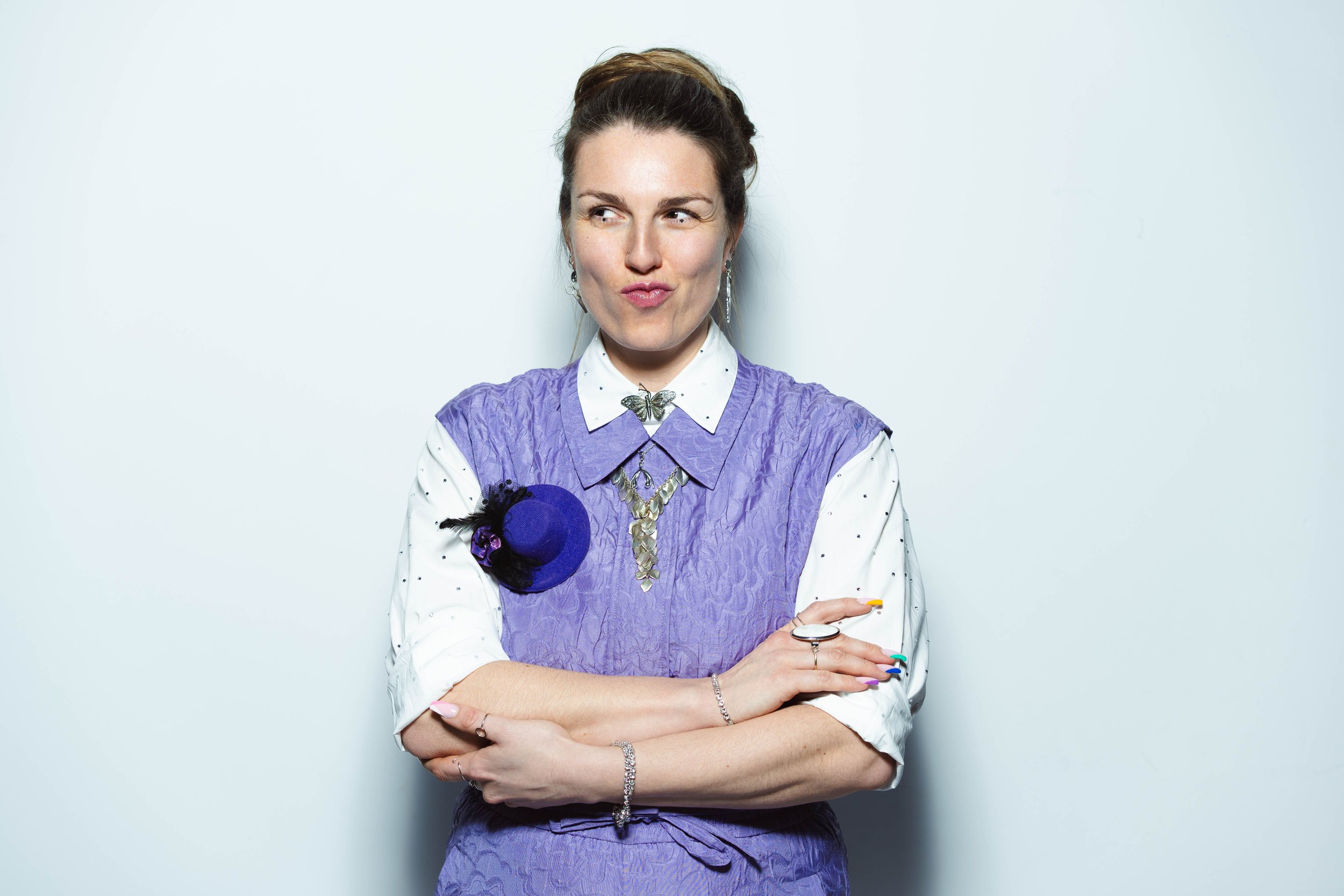 Vrouw met oprechte blik, gekleed in een paarse jurk en een witte blouse, met accessoires zoals oorringen, een ketting met een vlinder en een armband, staand met gekruiste armen voor een witte achtergrond.