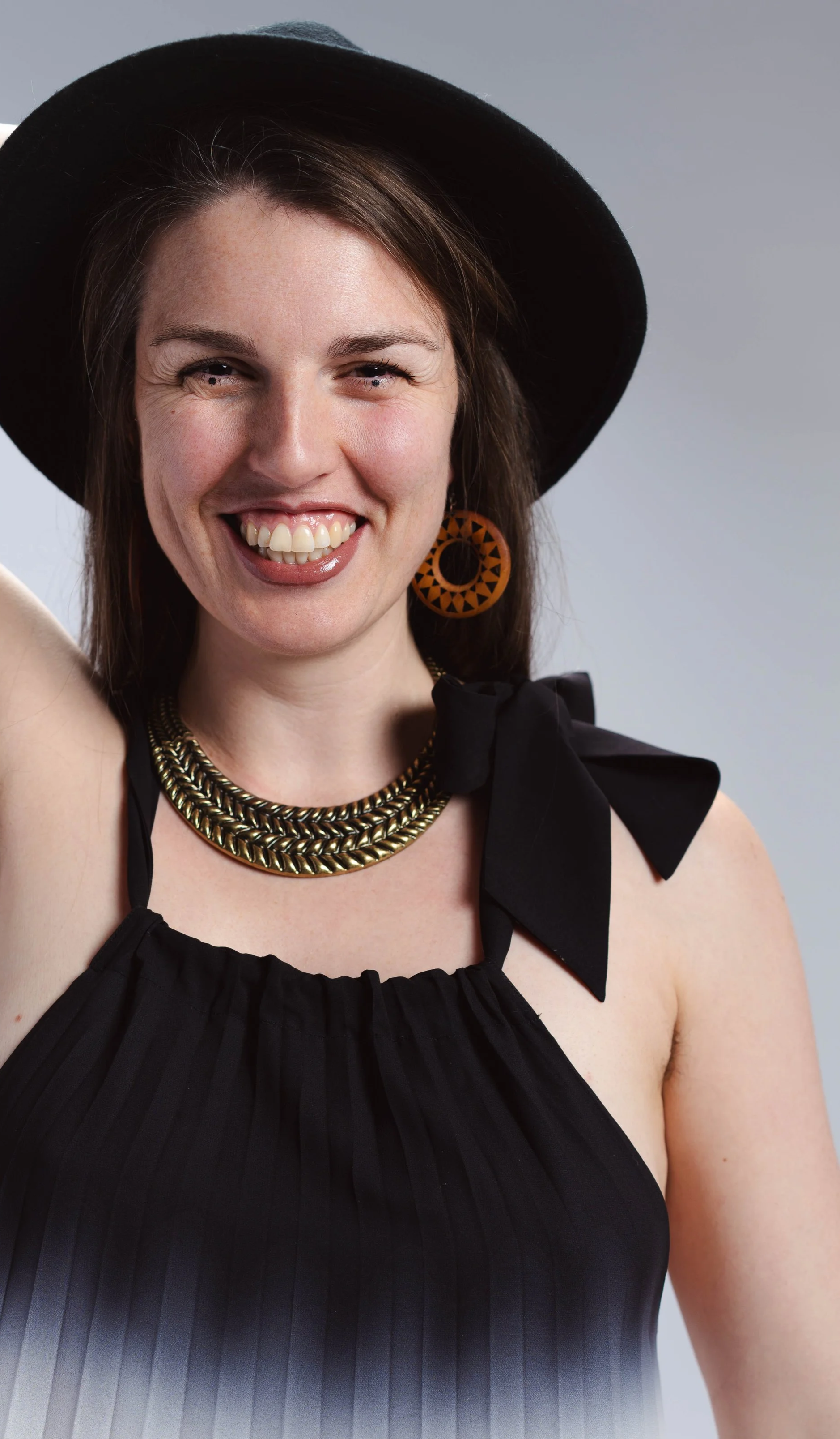 Een lachende vrouw in een zwarte jurk en hoed, met grote oorbellen sieraden, poseert tegen een neutrale achtergrond.