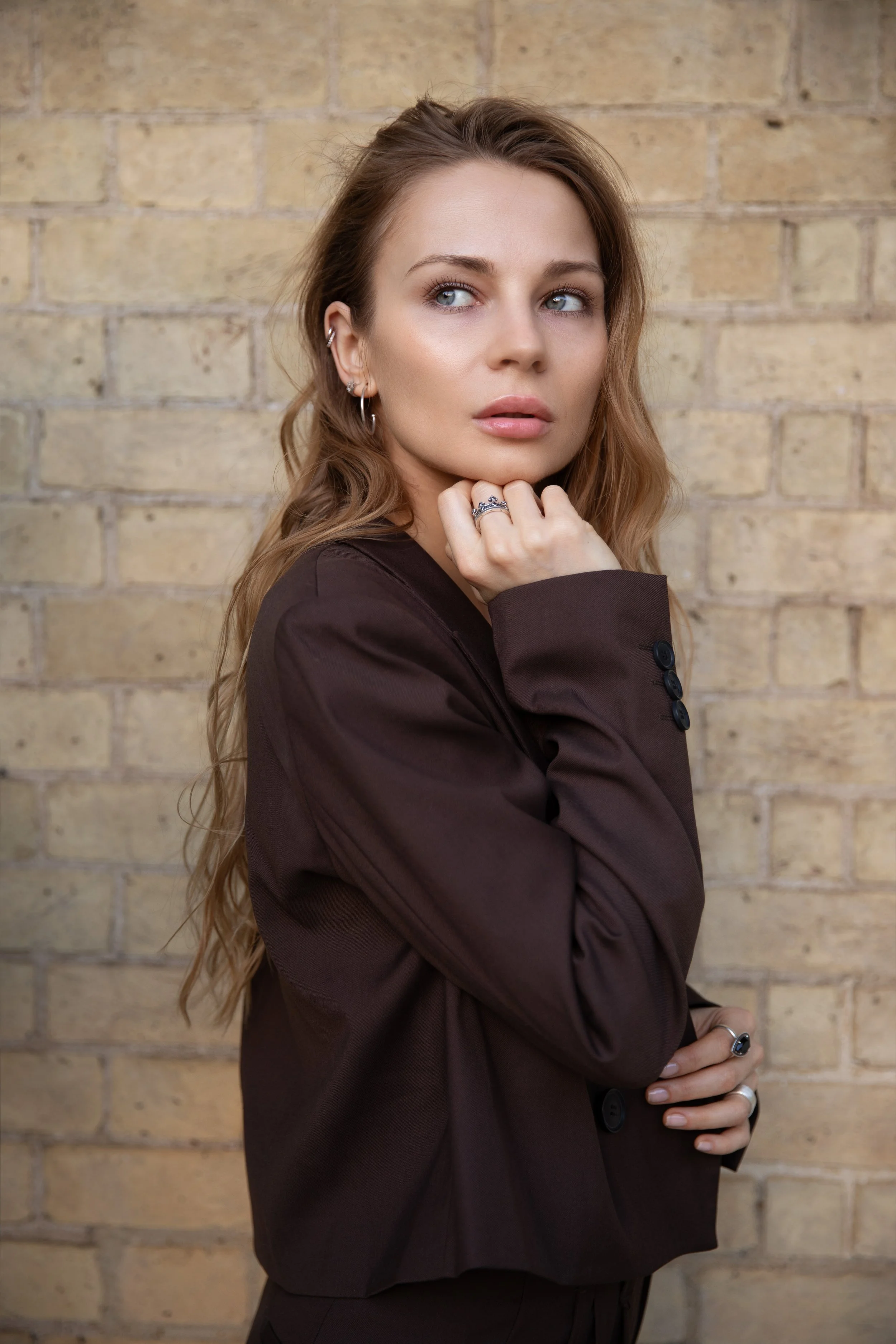 A woman with wavy hair looking away, wearing a dark blazer and jewelry, standing in front of a brick wall.
