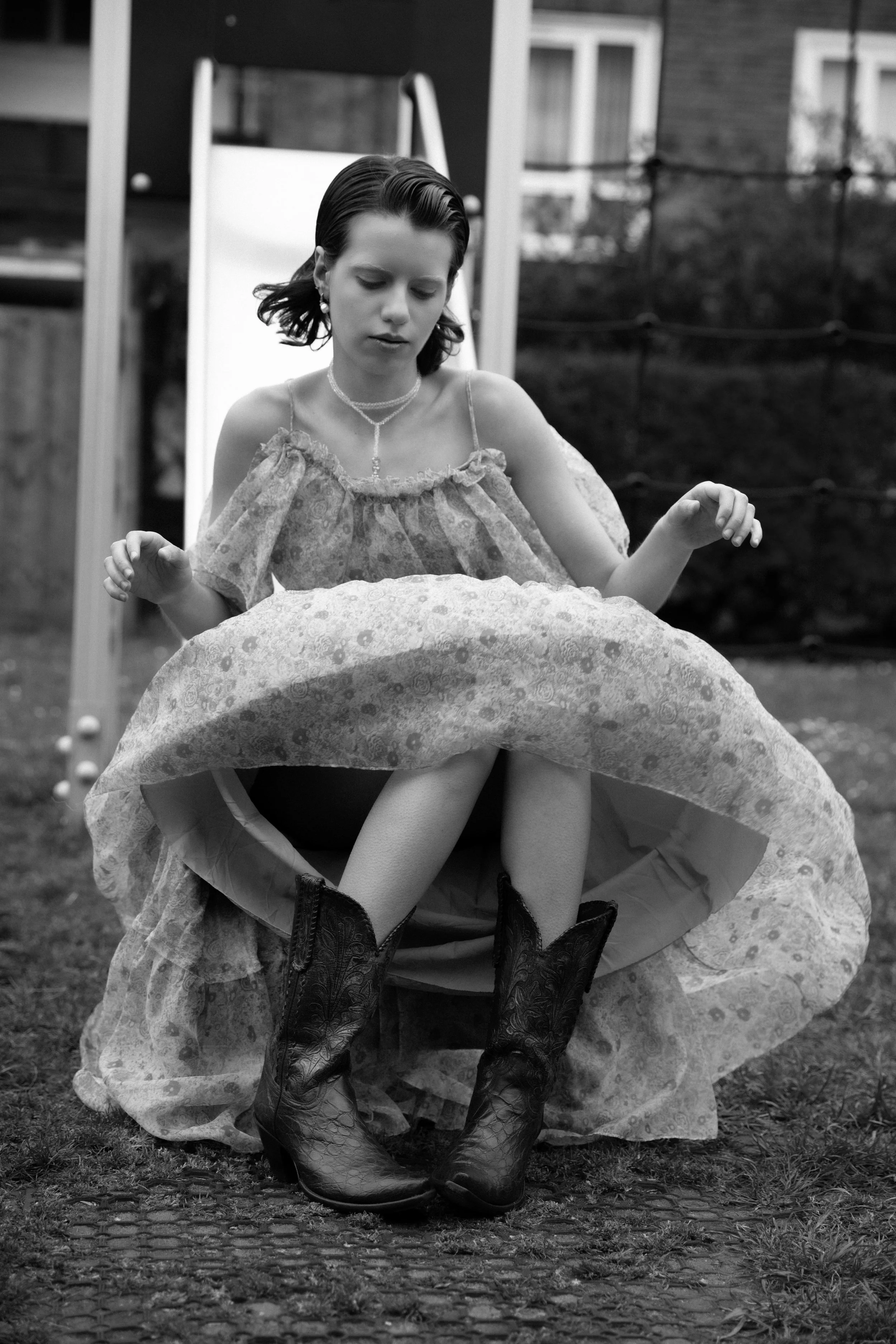 A woman sitting outdoors, lifting her floral skirt to show her black cowboy boots, with a slide in the background.