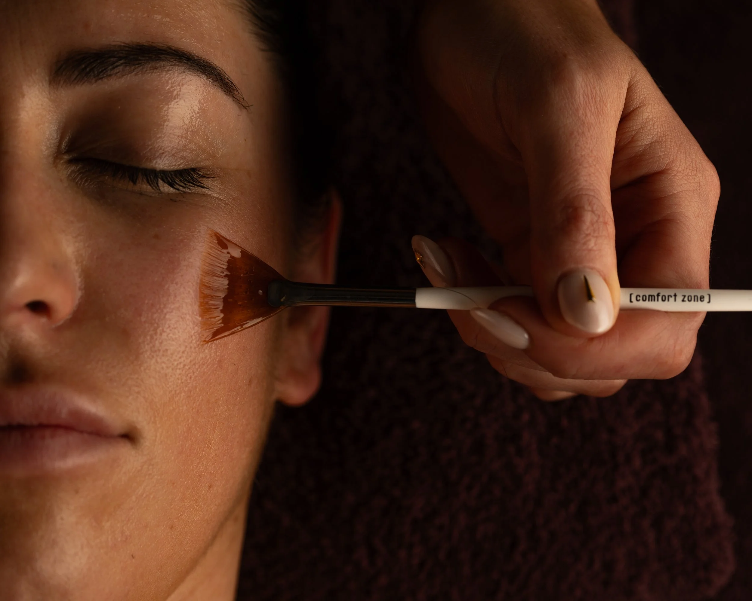 A person with closed eyes receives a facial treatment using a brush labeled 'comfort zone' on their cheek.