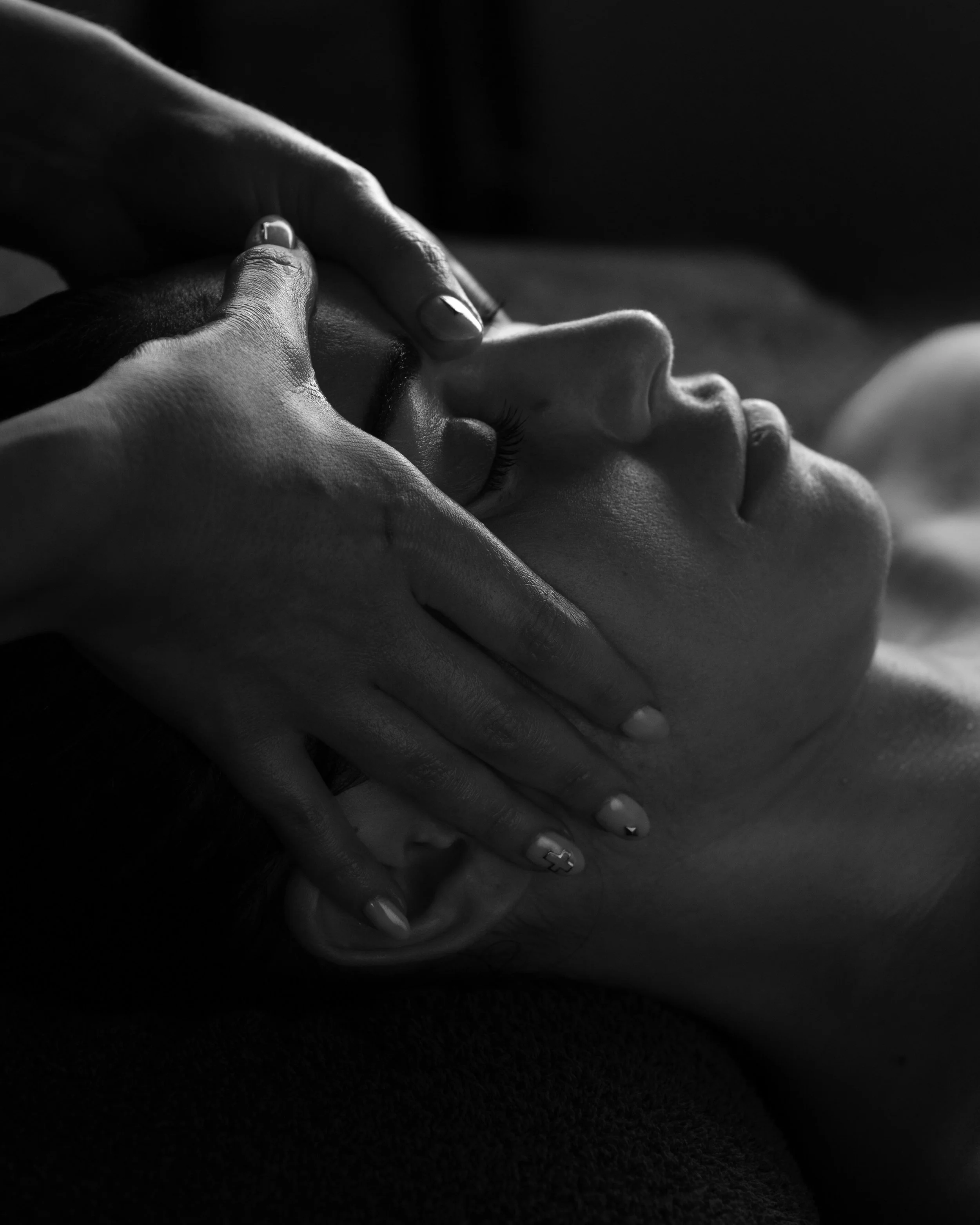 Close-up of a woman receiving a facial massage, lying down with eyes closed, in black and white.