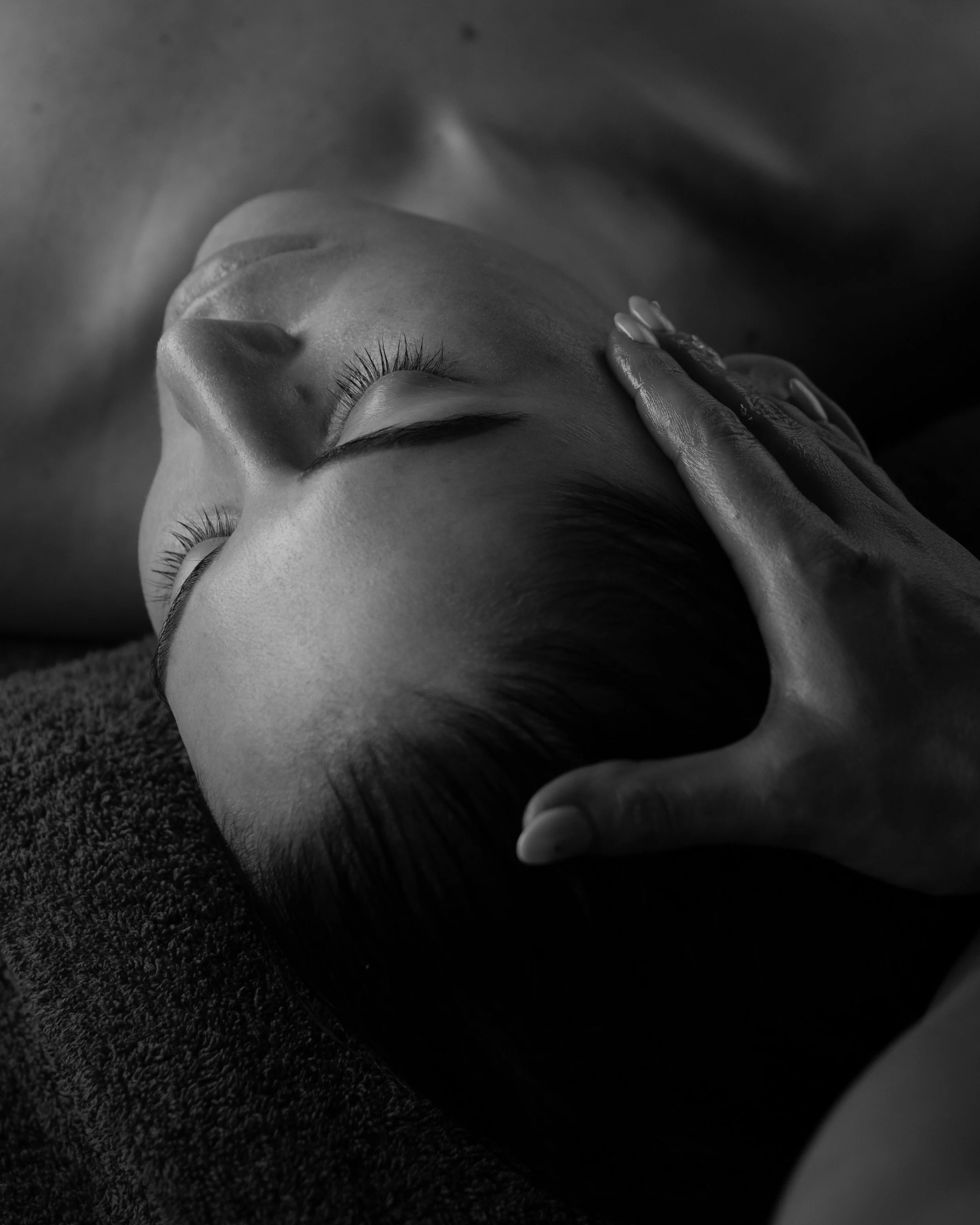 Close-up black and white photo of a person receiving a massage, lying on their back with eyes closed, head turned to the side, and hand gently placed on their temple.