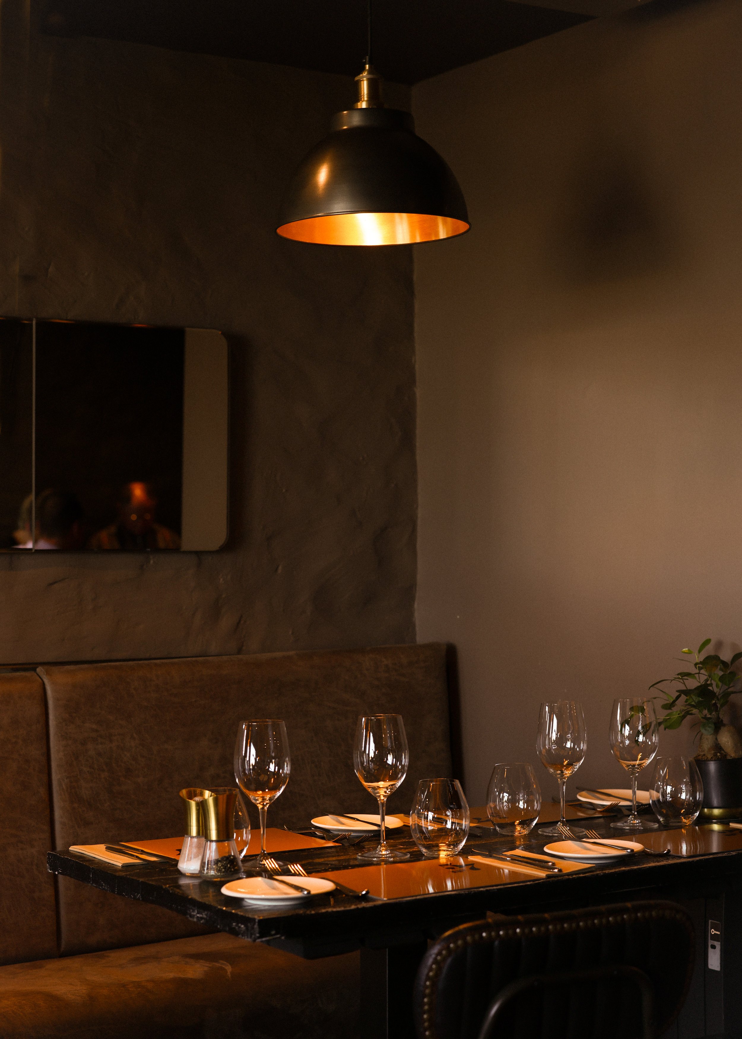 A dimly lit restaurant table set with empty wine glasses, plates, knives, and forks, with a dark wall and a black pendant light overhead.