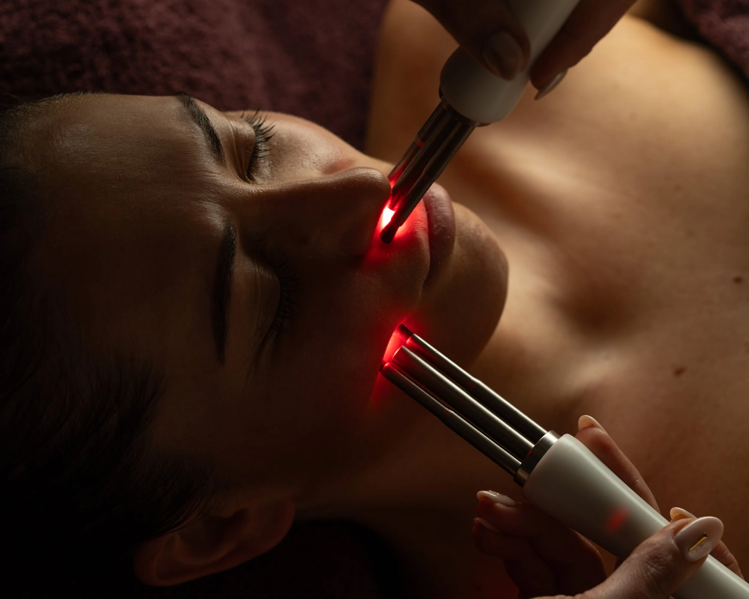 A woman receiving a facial treatment with a laser device directed at her face, in a spa or cosmetic clinic setting.