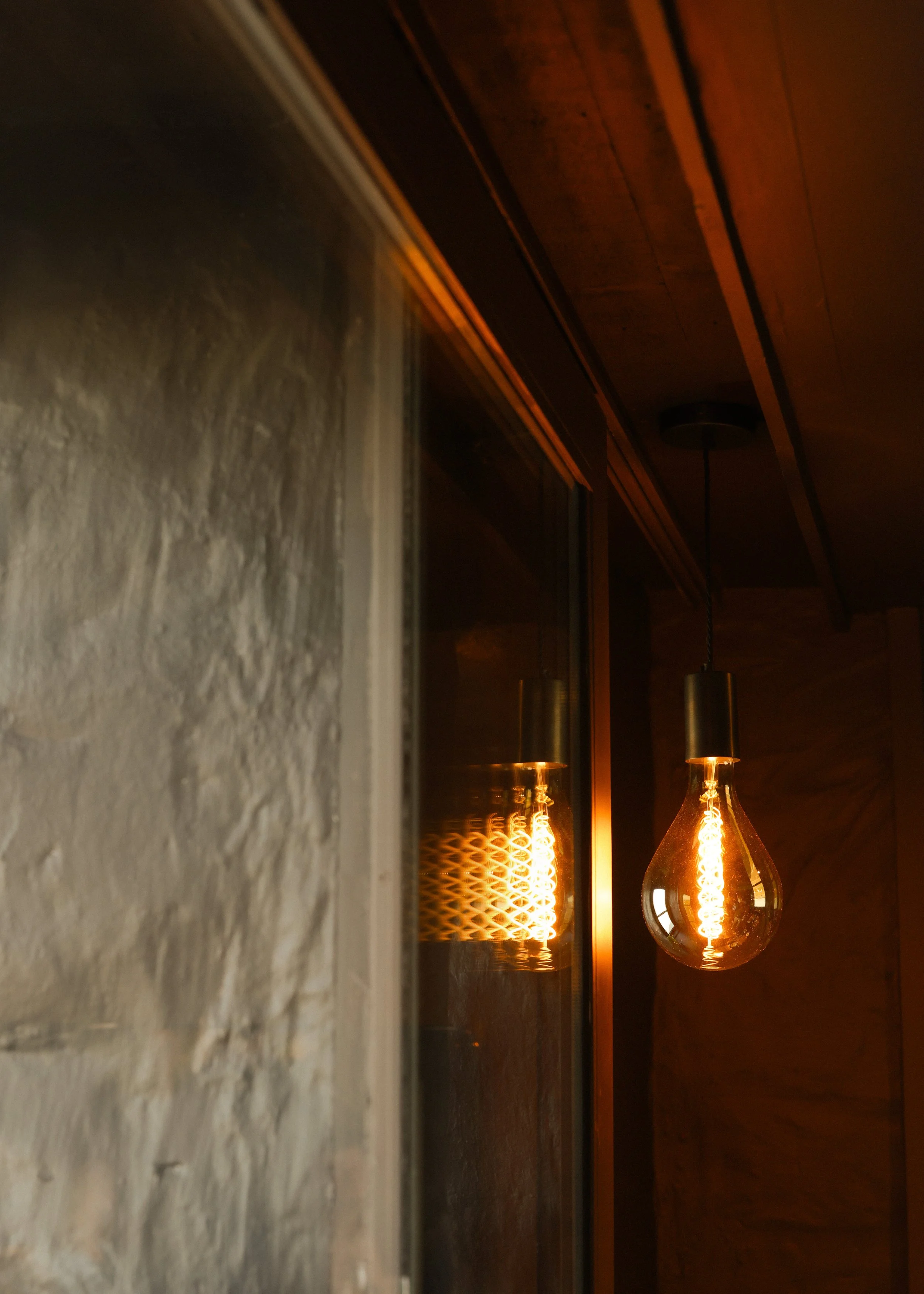 A lit filament light bulb hanging from a ceiling, reflecting in a mirrored surface on the wall, with a brick wall and wooden ceiling in the background.
