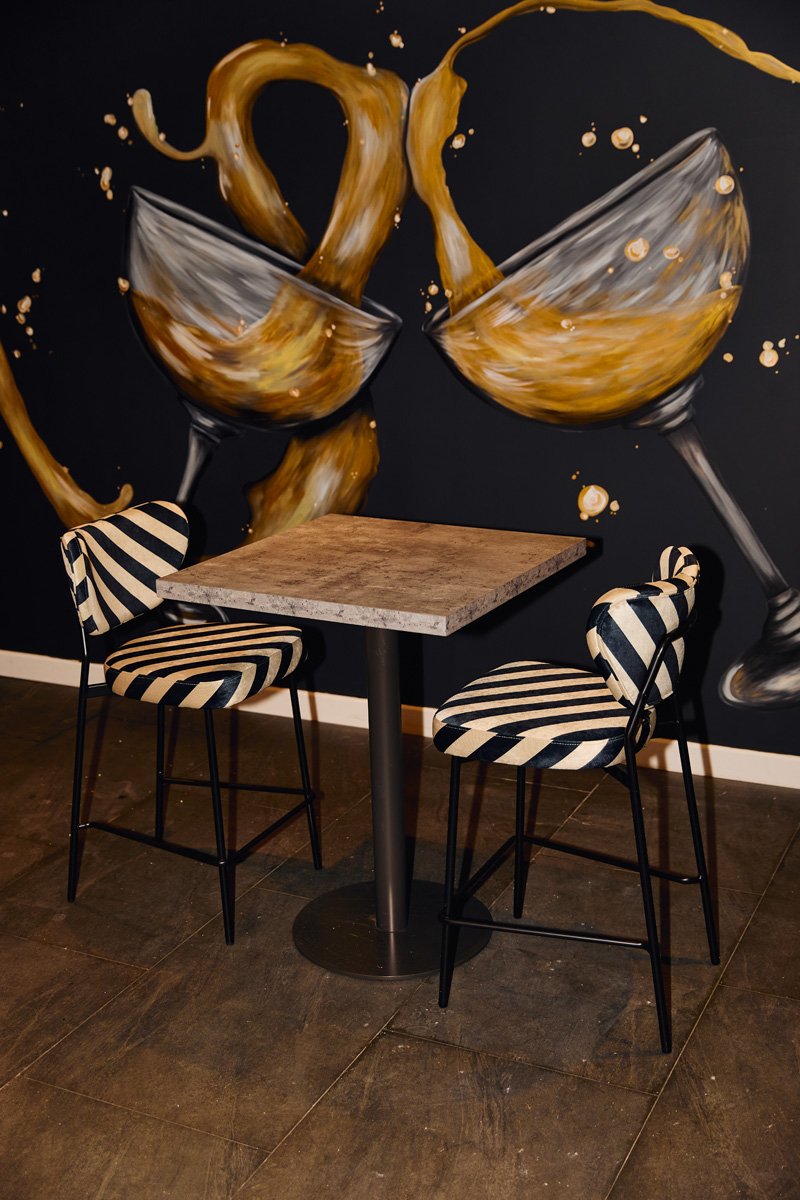 A small square table with a wood top and black metal base, surrounded by two chairs with black and beige striped upholstery and black metal legs, against a wall with a mural of wine glasses splashing with brown wine.