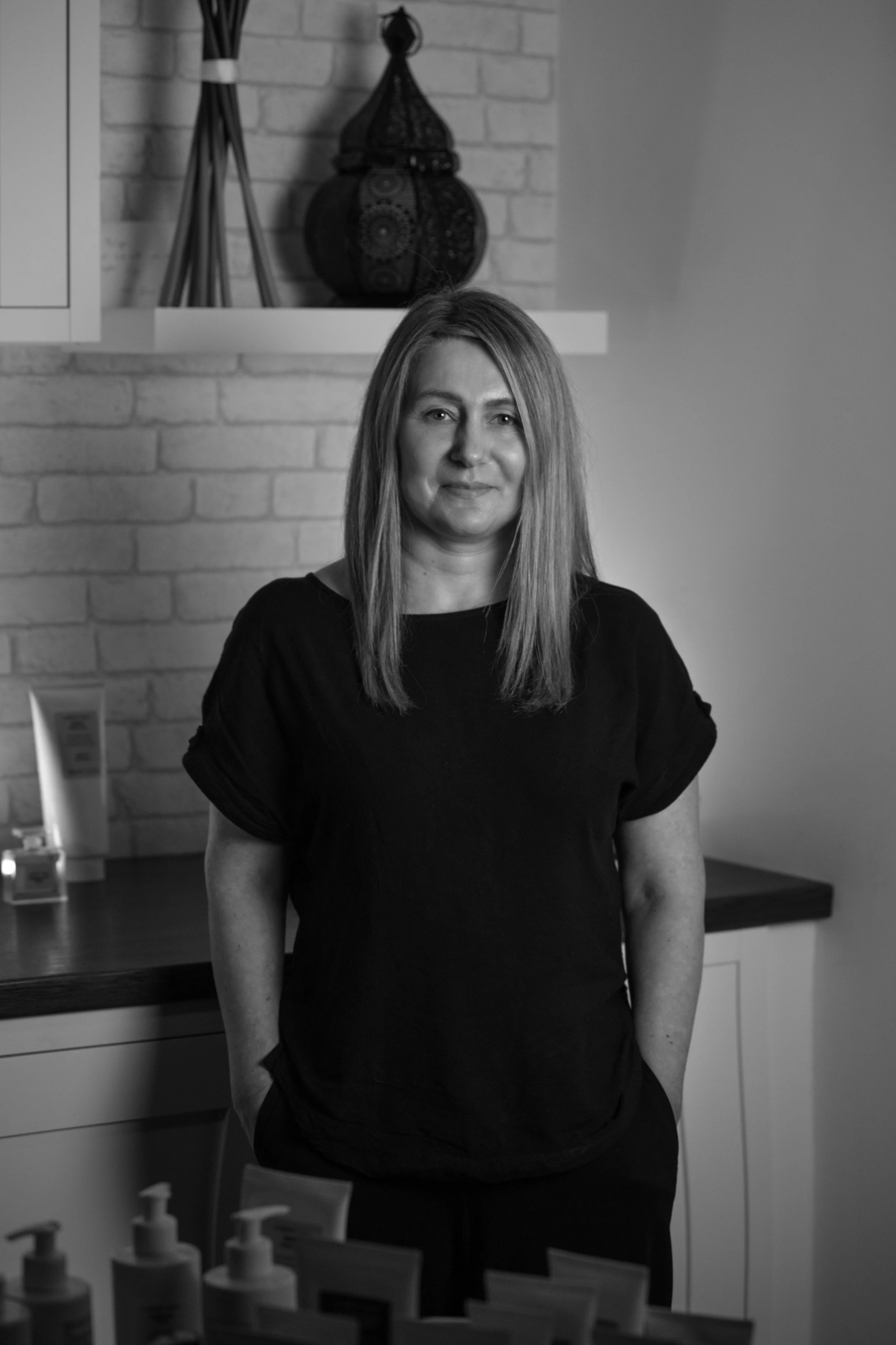 A woman with shoulder-length hair wearing a black top, standing indoors with bottles on a table in front of her and a brick wall with decorative objects behind her.