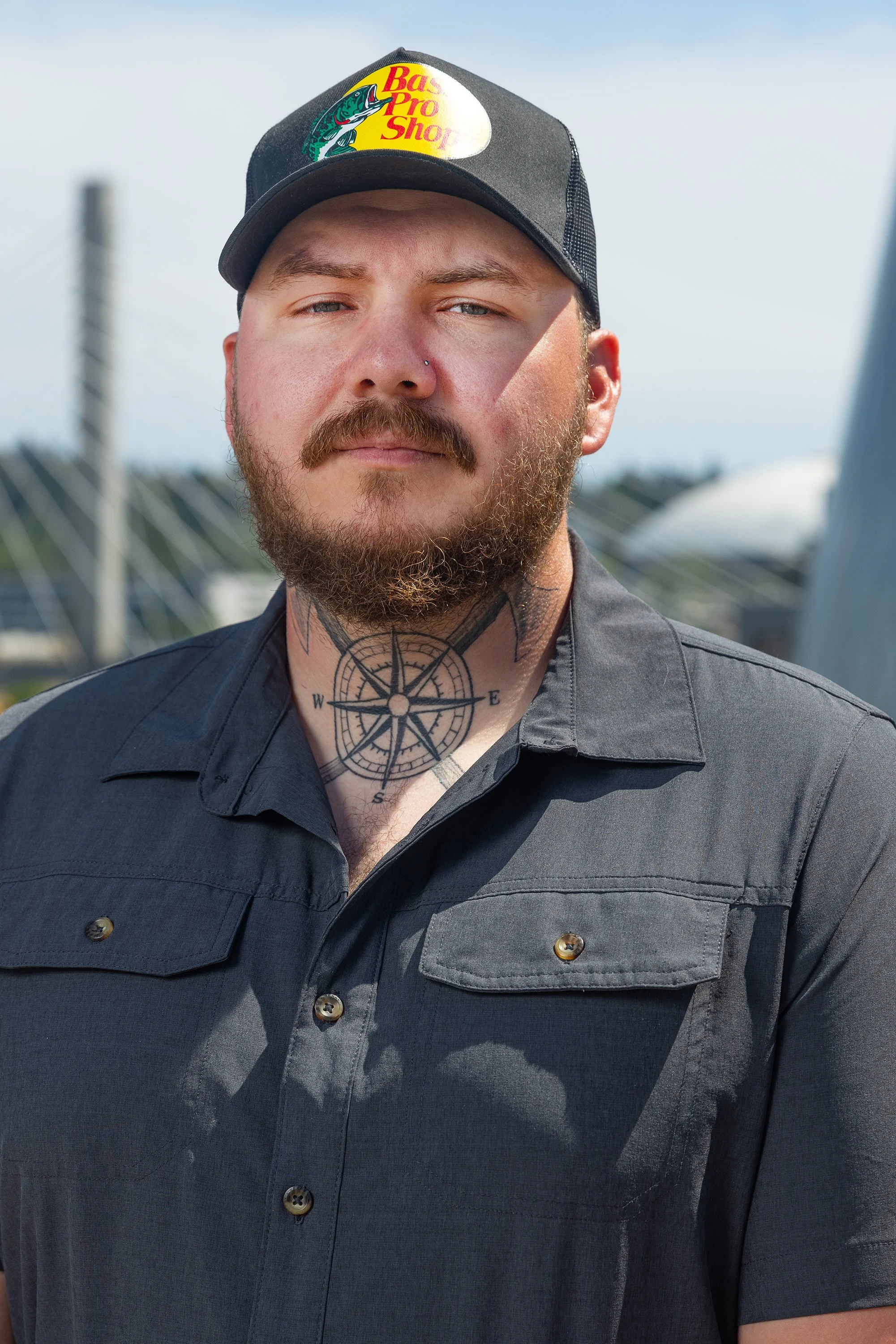 A close-up of a man with a beard and mustache, wearing a black cap with a Bass Pro Shops logo, a nose piercing, and a navy-blue button-up shirt outdoors with a blurry background of a bridge and sky.