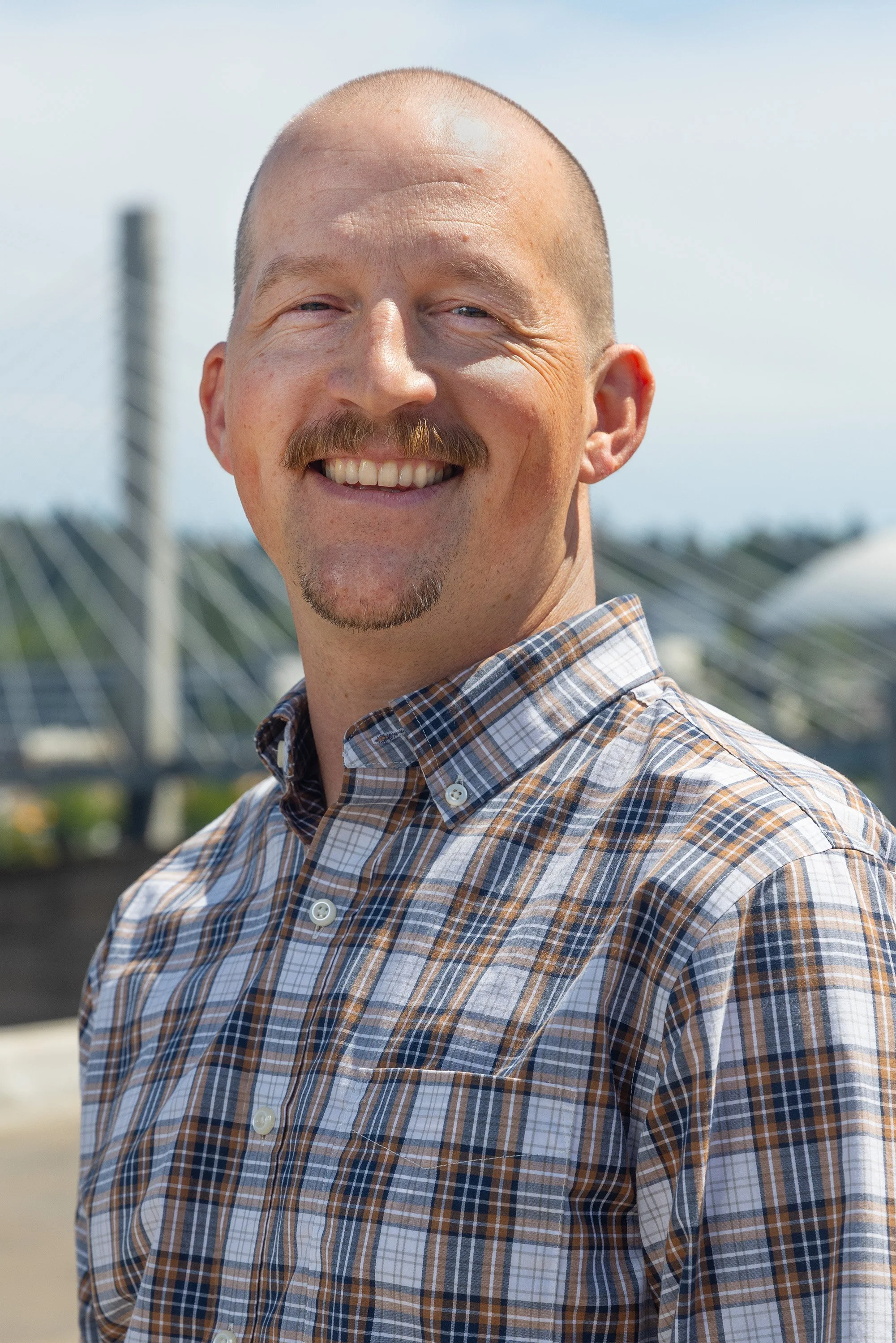 A smiling man with a bald head, light skin, and a short beard, wearing a plaid button-up shirt, standing outdoors during daytime with solar panels and a clear sky in the background.