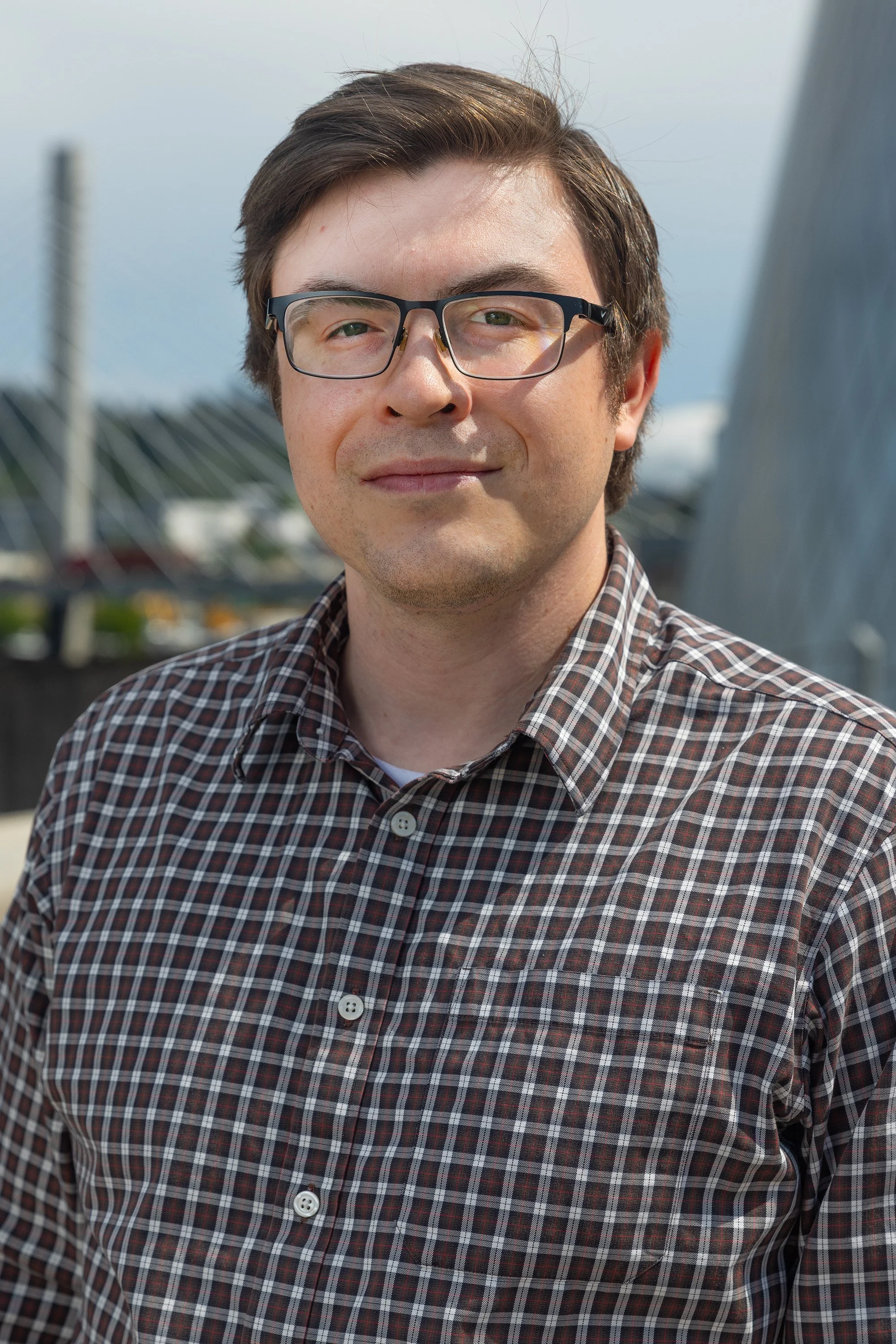 Portrait of a man with brown hair and glasses wearing a plaid shirt, outdoors with a modern bridge structure in the background.
