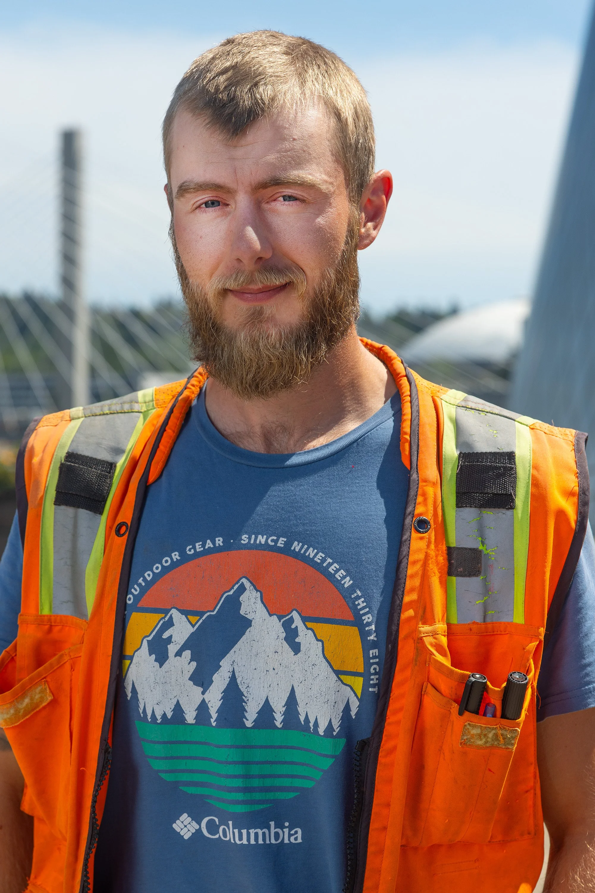 A man outdoors with a background of a bridge and a clear sky. He has a full beard and short hair, wearing a blue T-shirt with mountain graphic and the Columbia logo, and an orange safety vest with tools in the pocket.