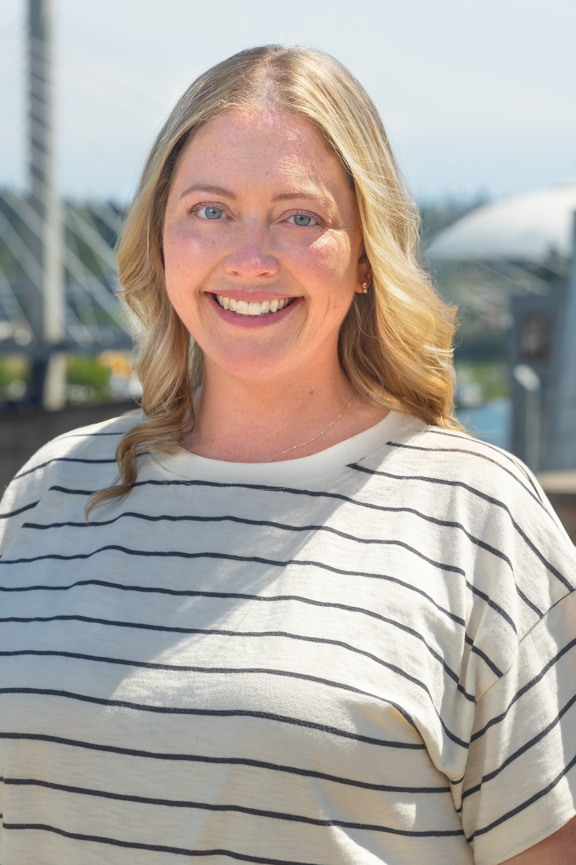 Close-up of a woman with blonde hair and blue eyes smiling outdoors in sunlight, wearing a white and navy striped shirt.