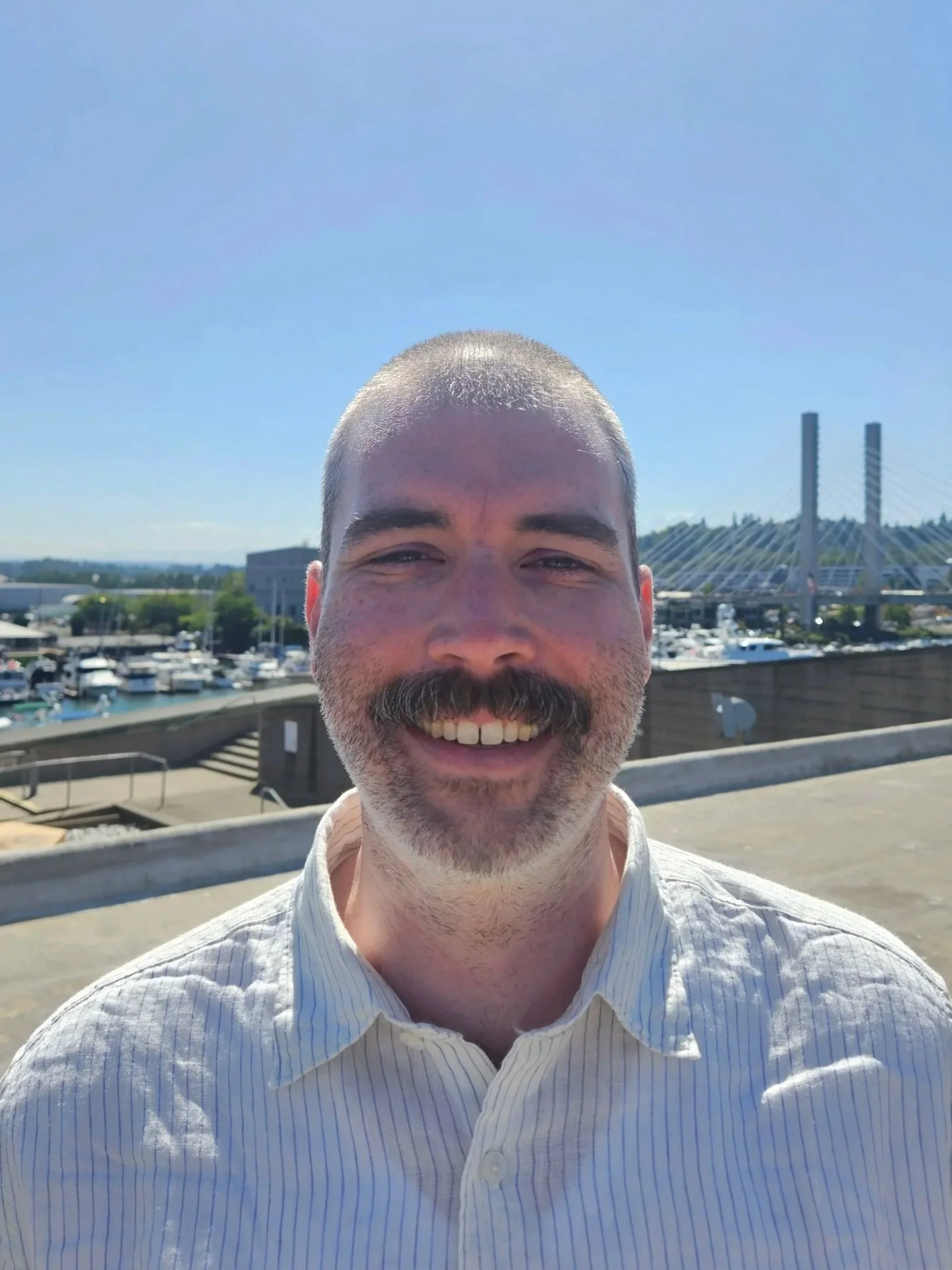 A smiling man with a beard and short hair, wearing a light-colored striped shirt, standing outdoors near a harbor with boats and a bridge in the background on a sunny day.