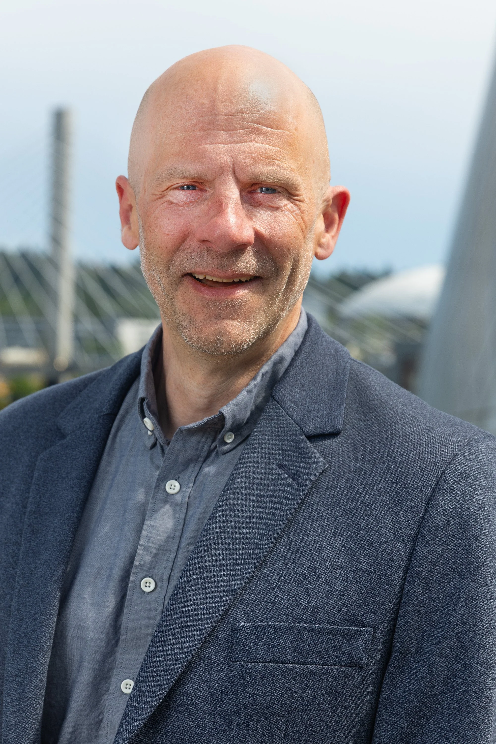 Close-up of a bald man with a gray beard, wearing a dark gray blazer and a gray collared shirt, standing outdoors with a blurred bridge and cloudy sky in the background.