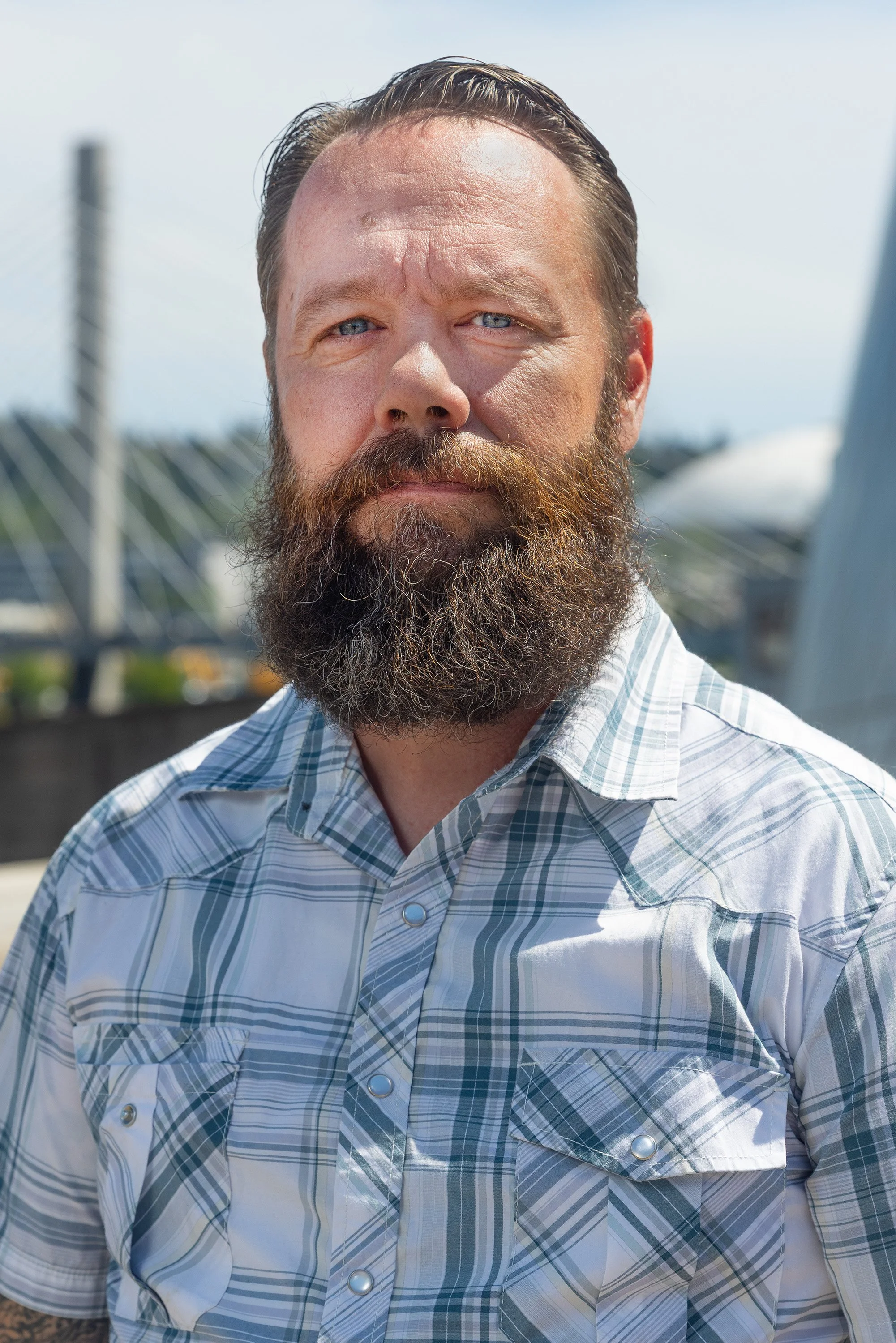 A man with a full beard and slicked-back hair wearing a blue and white striped button-up shirt standing outdoors on a sunny day.