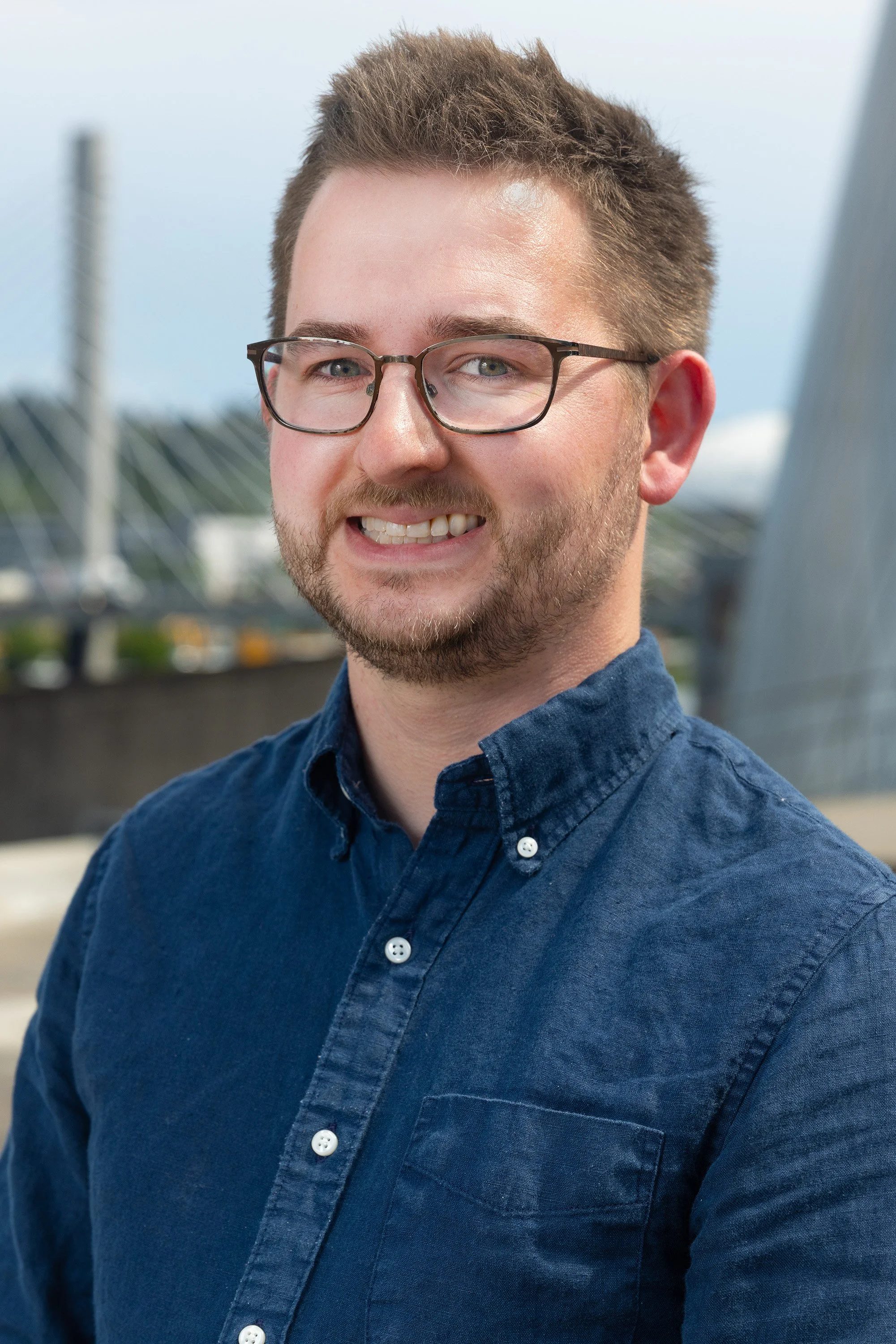 Portrait of a young man with glasses, short brown hair, a beard, wearing a blue button-up shirt, smiling outdoors with a bridge and cloudy sky in the background.