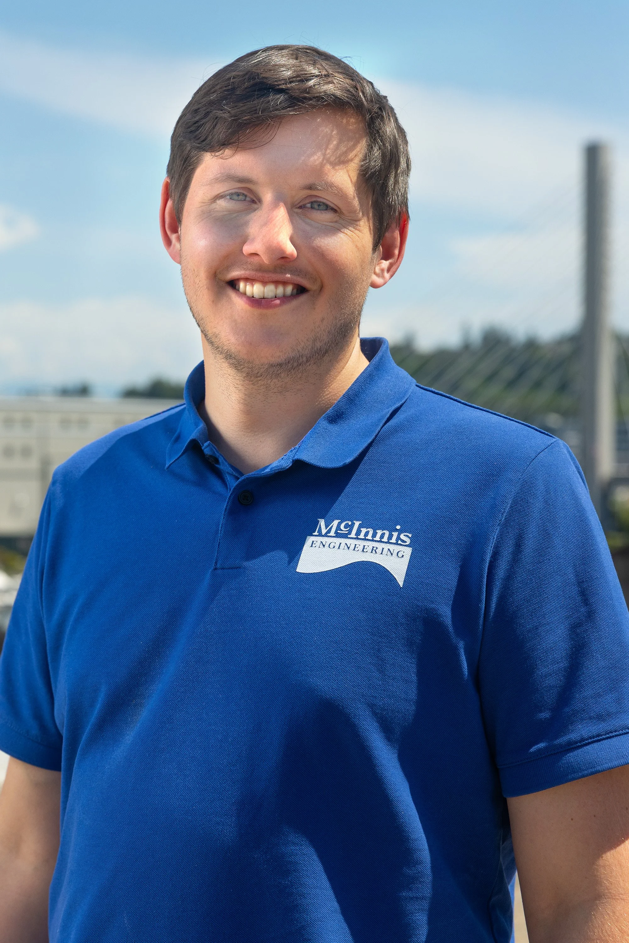 A young man with light brown hair and blue eyes smiling outdoors, wearing a blue polo shirt with a McInnis Engineering logo, with a bridge and clear sky in the background.