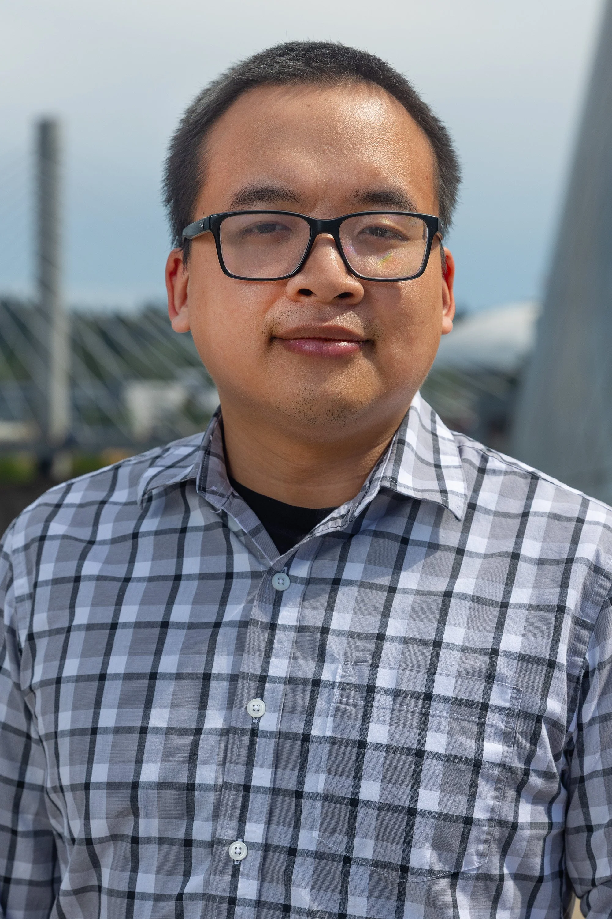 A man wearing glasses and a checkered shirt standing outdoors with a cloudy sky and a large metallic structure in the background.