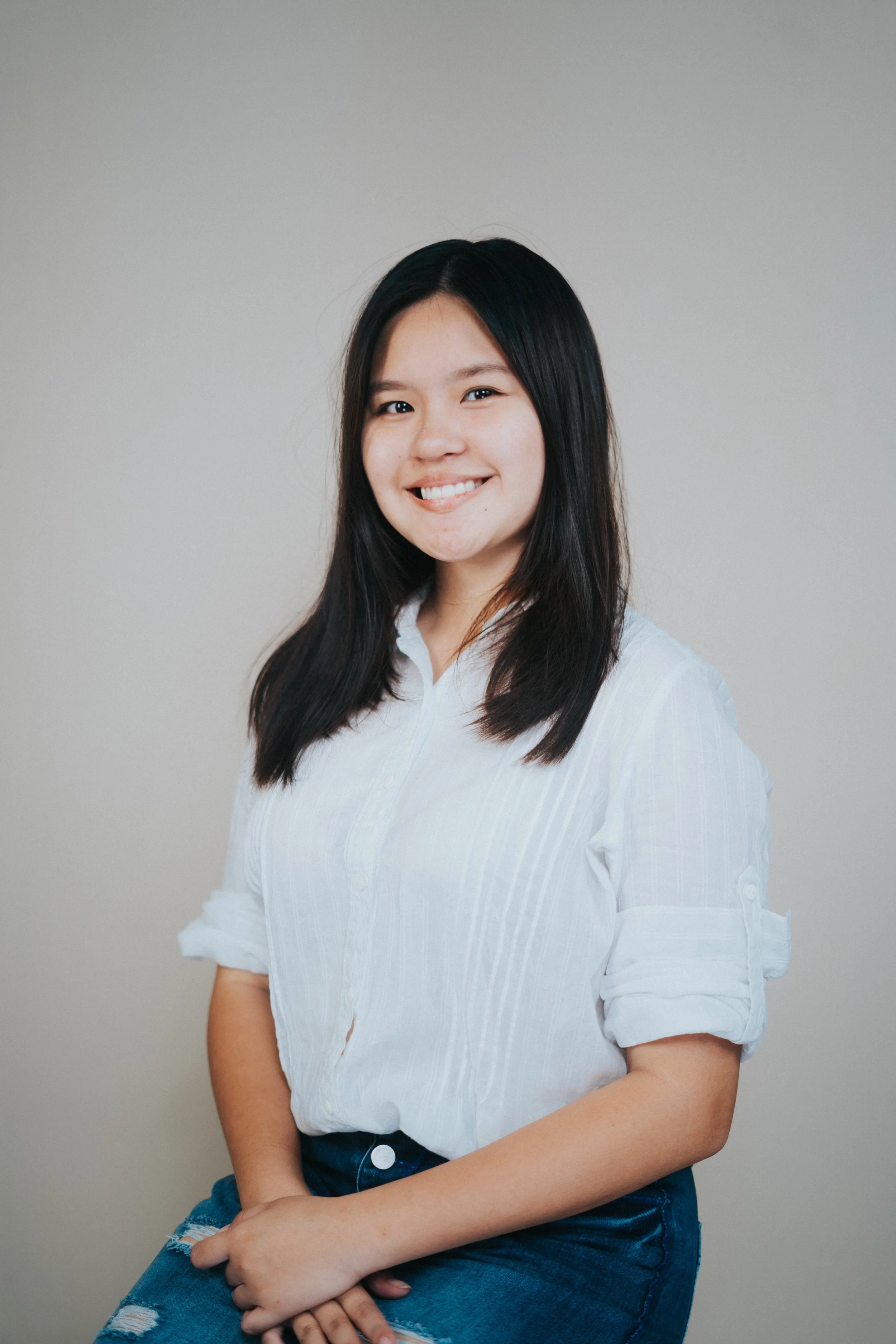 Young woman with long black hair smiling, wearing a white blouse with rolled-up sleeves and distressed jeans, sitting with crossed hands against a plain gray background.