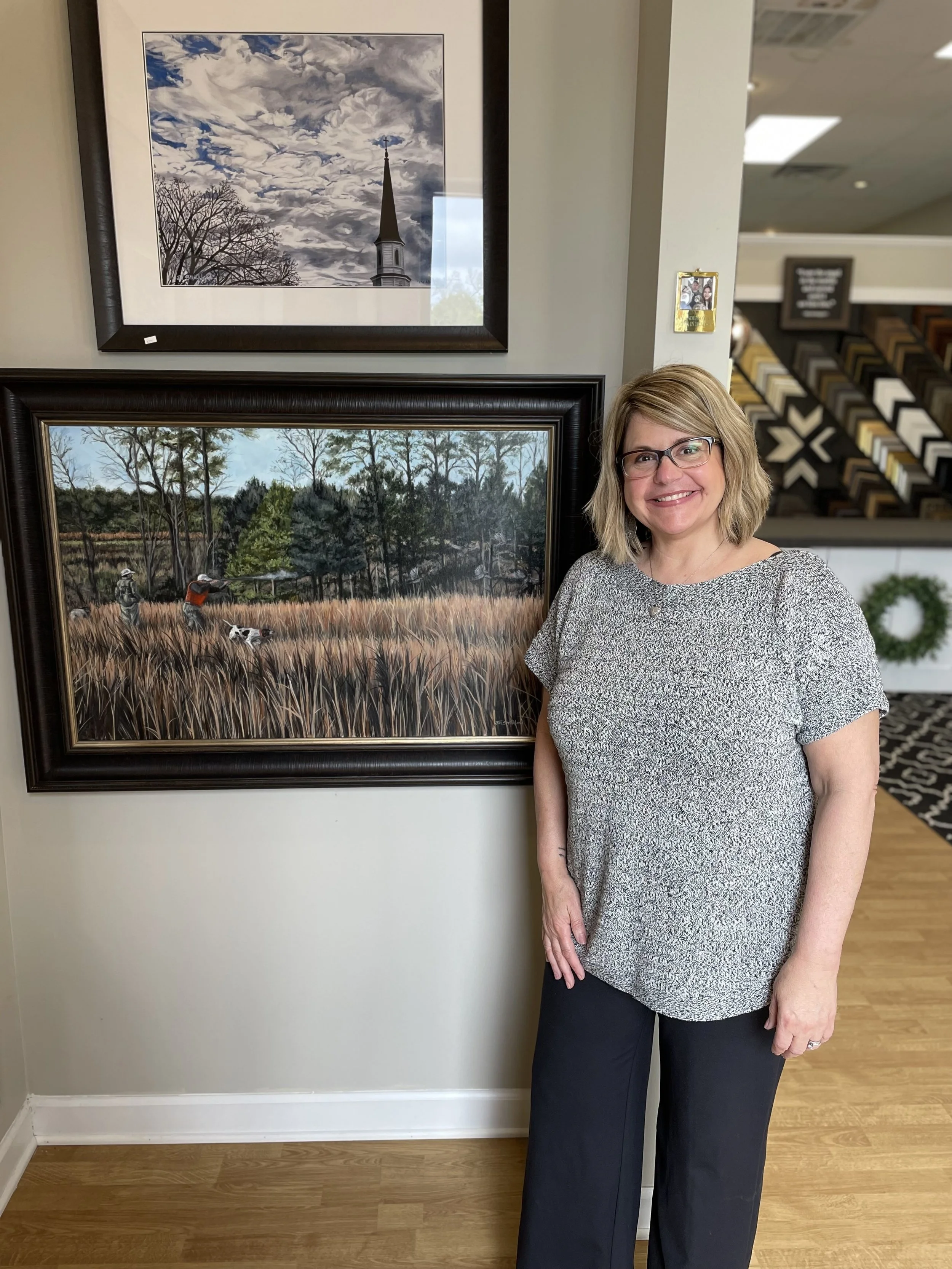 A woman with glasses and blonde hair smiling, standing indoors next to two framed landscape paintings, one above her and one beside her. The background includes a display of picture frames and a carpeted area with patterned flooring.