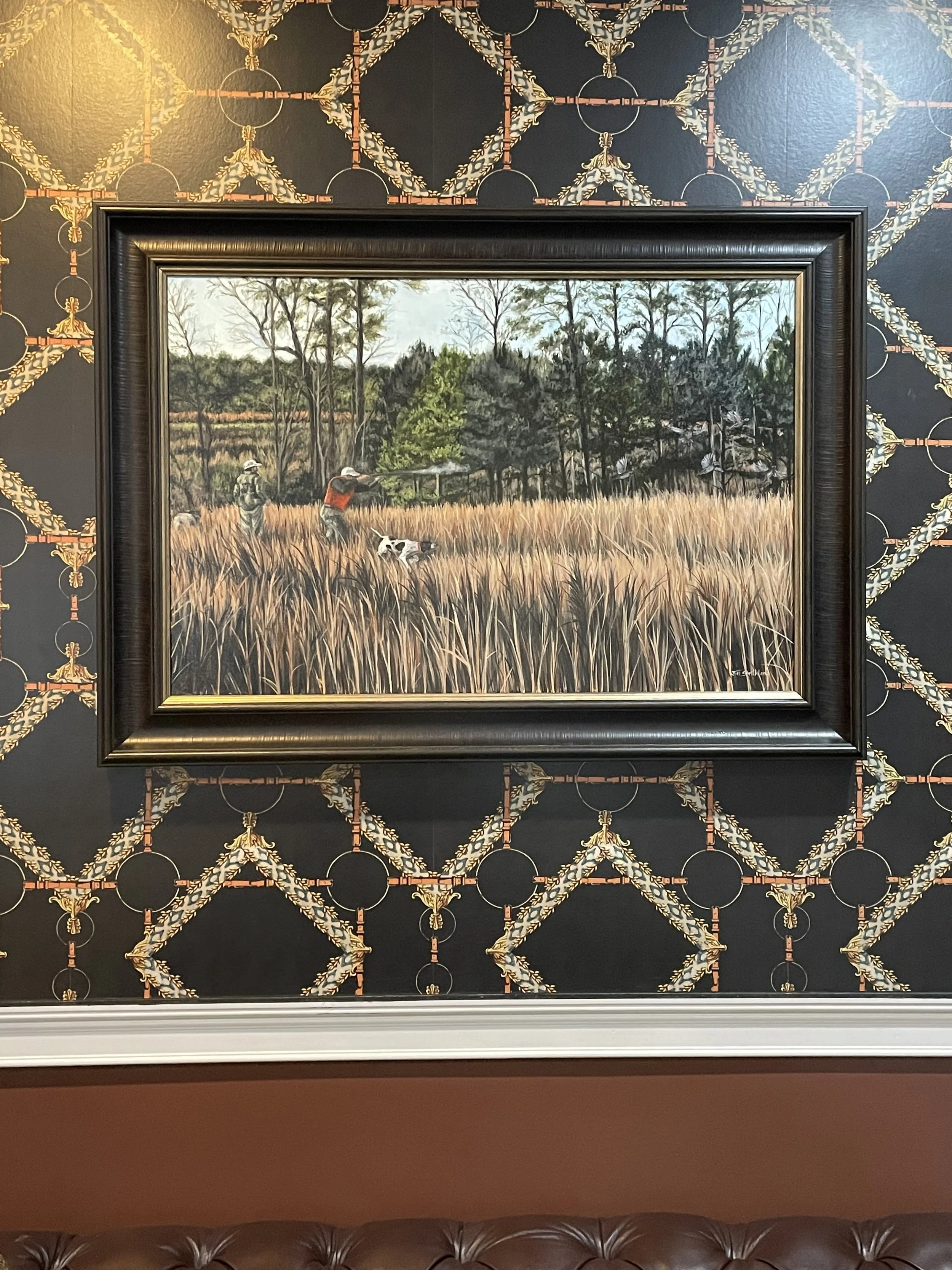 Painting of two hunters and a dog in a field of tall grass with trees in the background, displayed on a wall with decorative black, gold, and red patterned wallpaper.