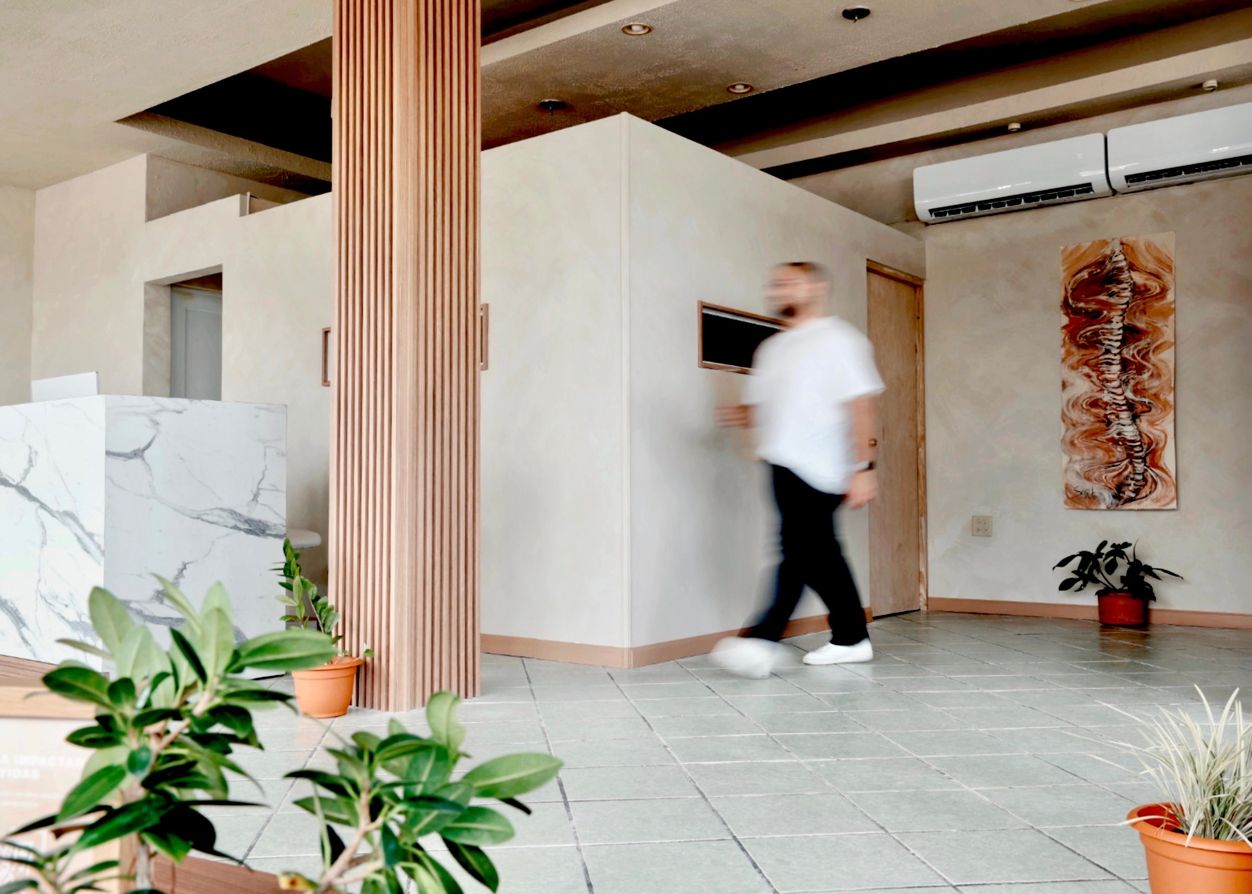 A man in white shirt and black pants walking through a modern, minimalist interior with potted plants, abstract art on the wall, and wooden accents.