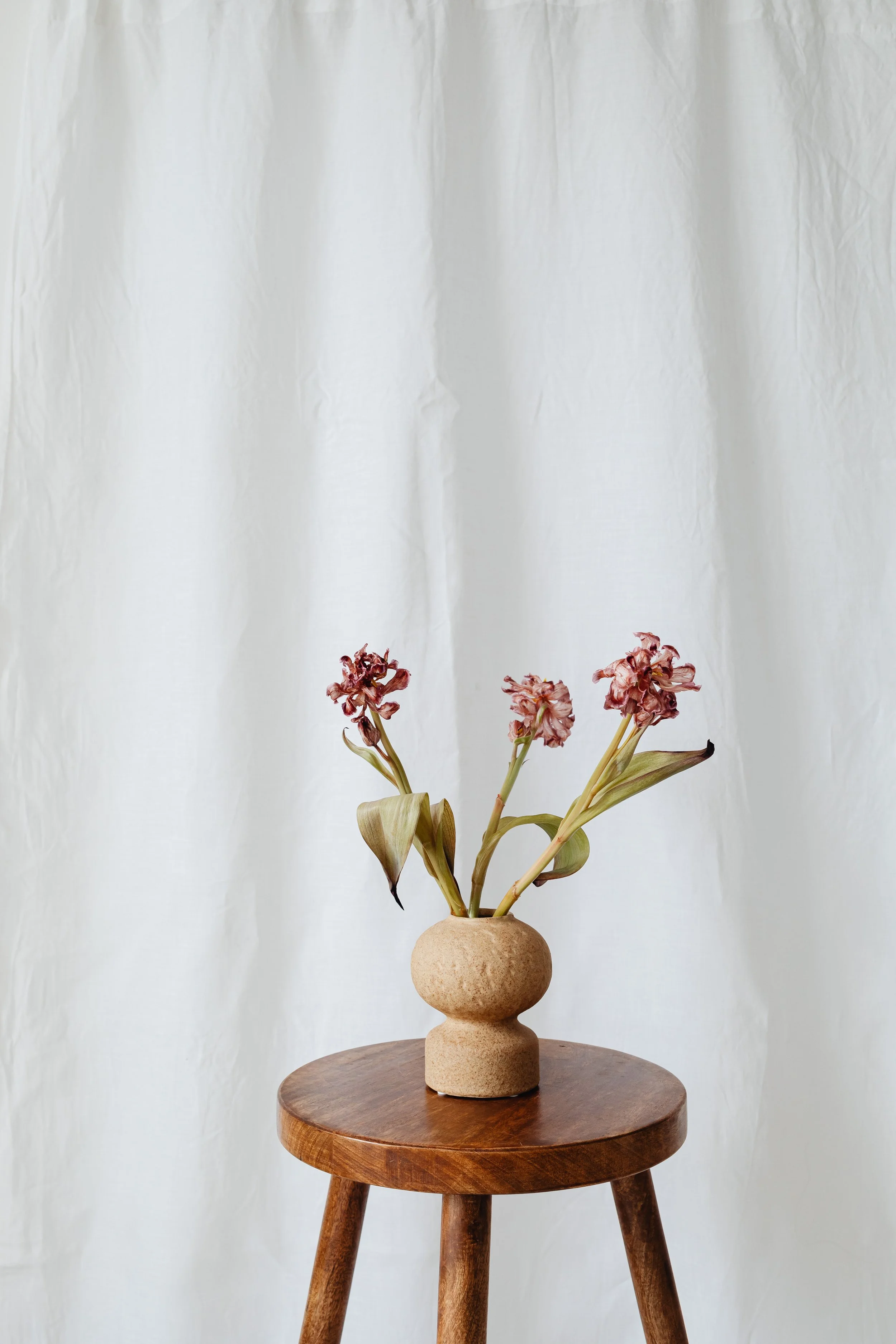 A beige, textured, abstract vase on a round wooden table with three legs, holding wilted pink flowers with green leaves against a plain white backdrop.