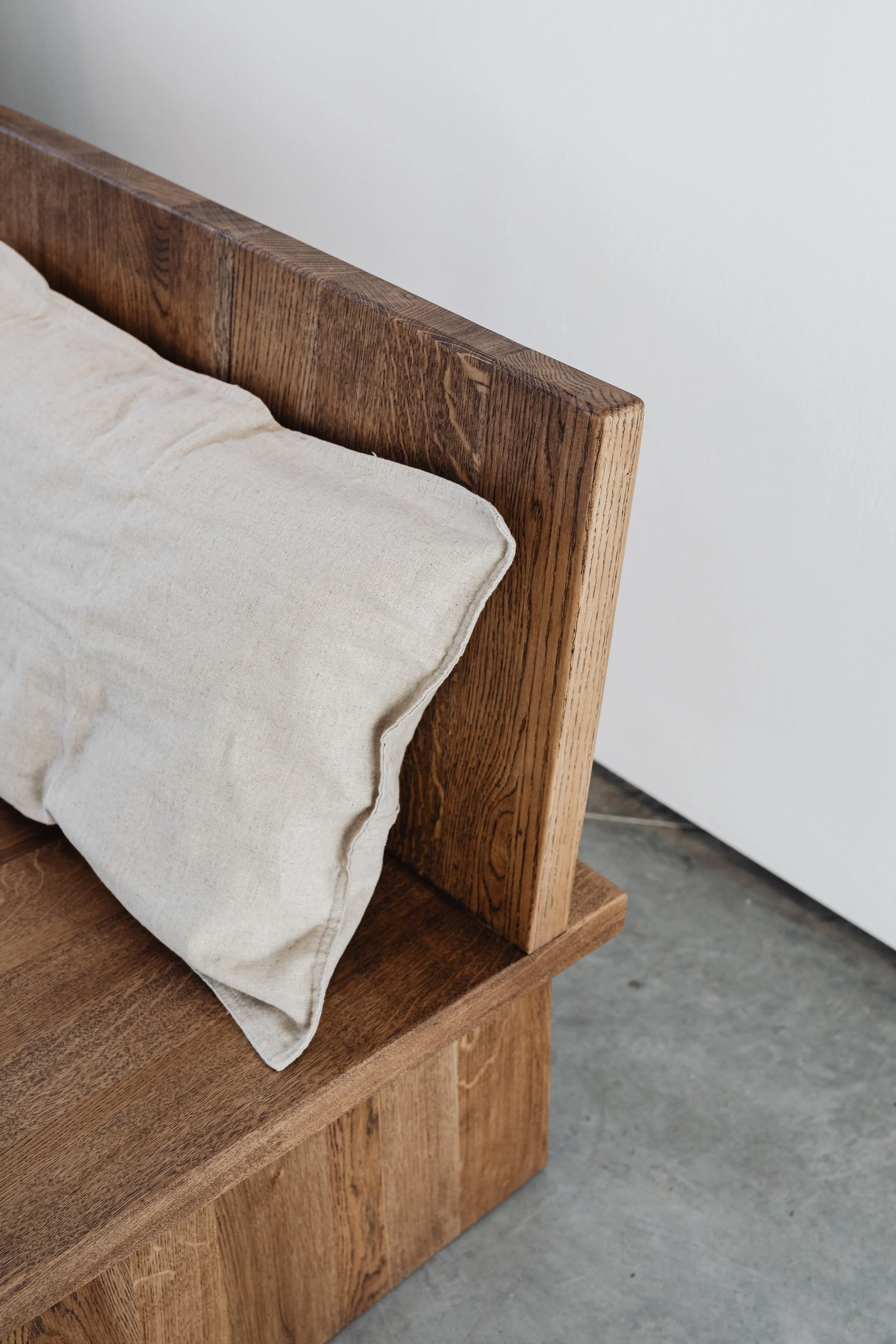 Close-up of a wooden bed frame with a beige pillow on a wooden bed surface, against a plain white wall.