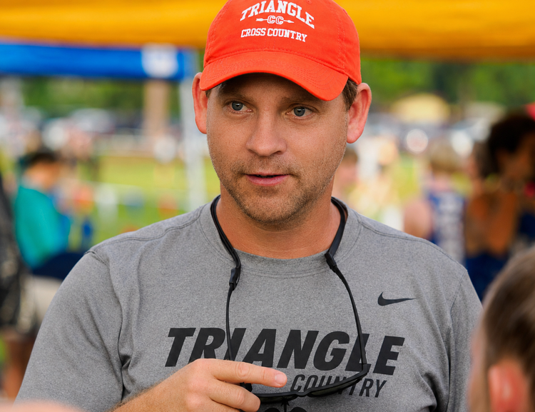 A man wearing a red cap and a gray T-shirt with 'TRIANGLE CROSS COUNTRY' on it, standing outdoors at a cross-country event.