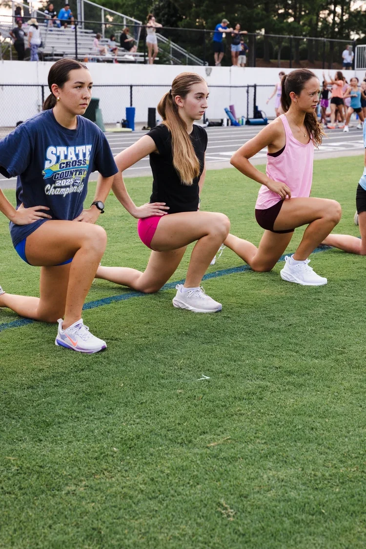 Three young women stretching on a football field while wearing athletic clothing, with other athletes and spectators in the background.