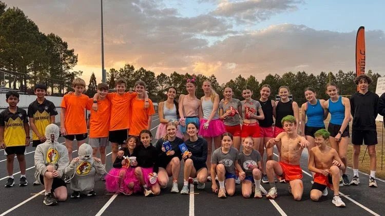 Group of children and teenagers posing on a track field during a sports event, some dressed in athletic clothing, others in costumes, with a sunset sky in the background.