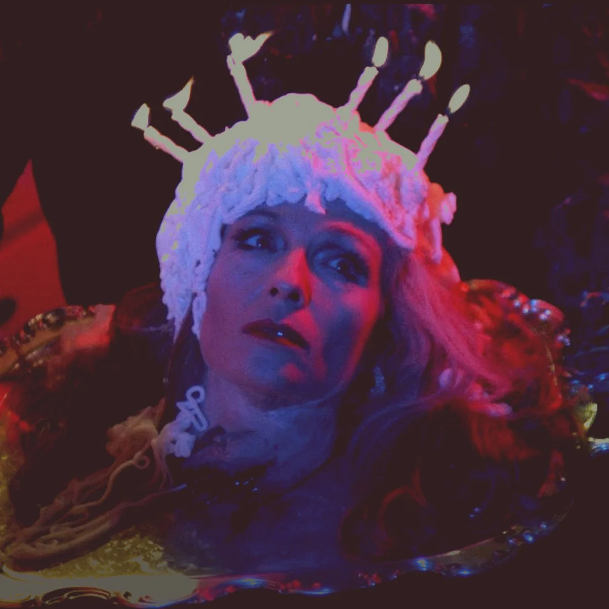 A woman with bright red hair and dark makeup is wearing a headpiece that resembles a crown made of candles. The lighting in the scene is dark with red and purple tones, giving a dramatic and theatrical appearance.