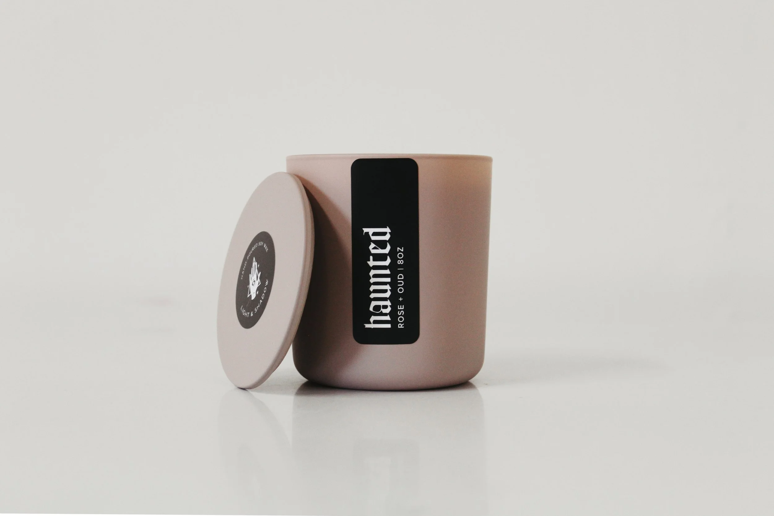 Pink container with a black label reading 'Haunted Rose + Oud 8oz' and a matching round lid with a black and white label.
