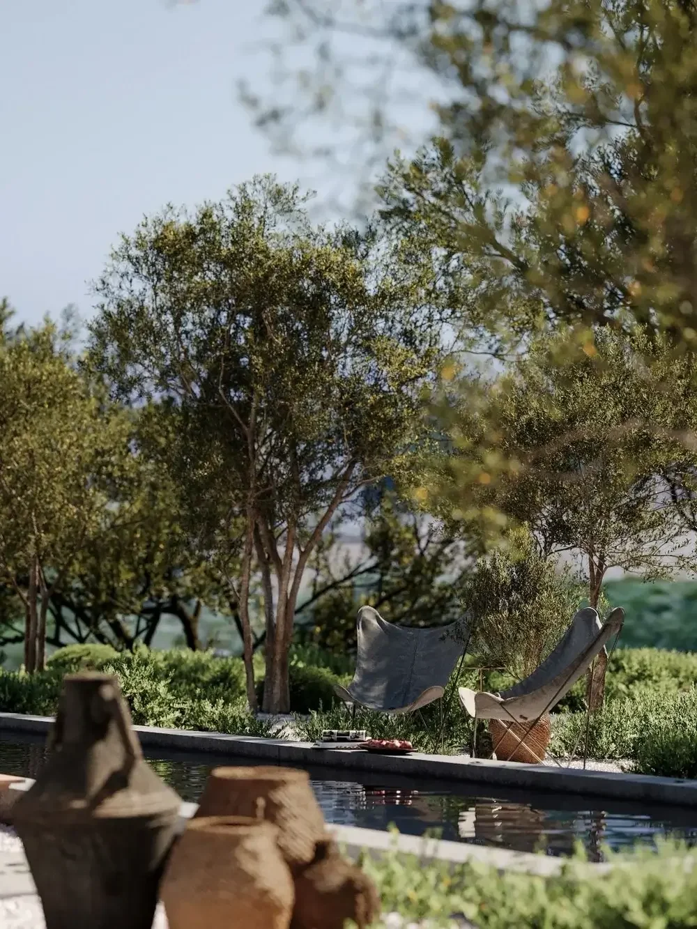 A peaceful outdoor scene with two chairs under trees, a small table between them, a swimming pool, and pottery in the foreground.