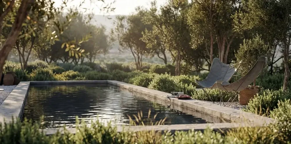 A peaceful outdoor scene featuring a rectangular swimming pool surrounded by lush green bushes and trees, with two modern lounge chairs and a basket nearby, bathed in warm sunlight.