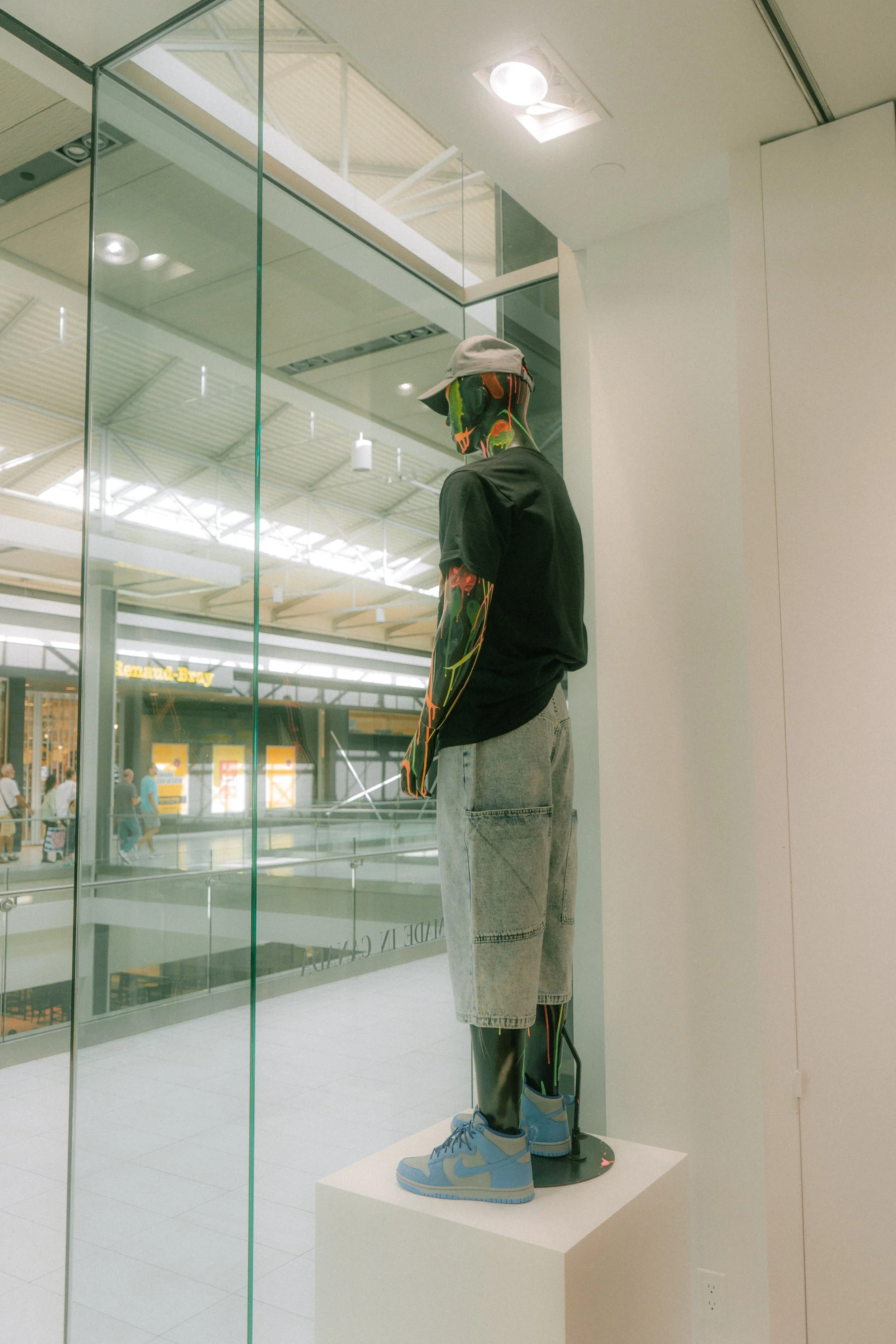 A mannequin displayed in a store wearing a black T-shirt, light denim shorts, and blue sneakers. The mannequin's head and arms have a colorful design. The setting appears to be a shopping mall with a glass wall separating the store from the walkway.