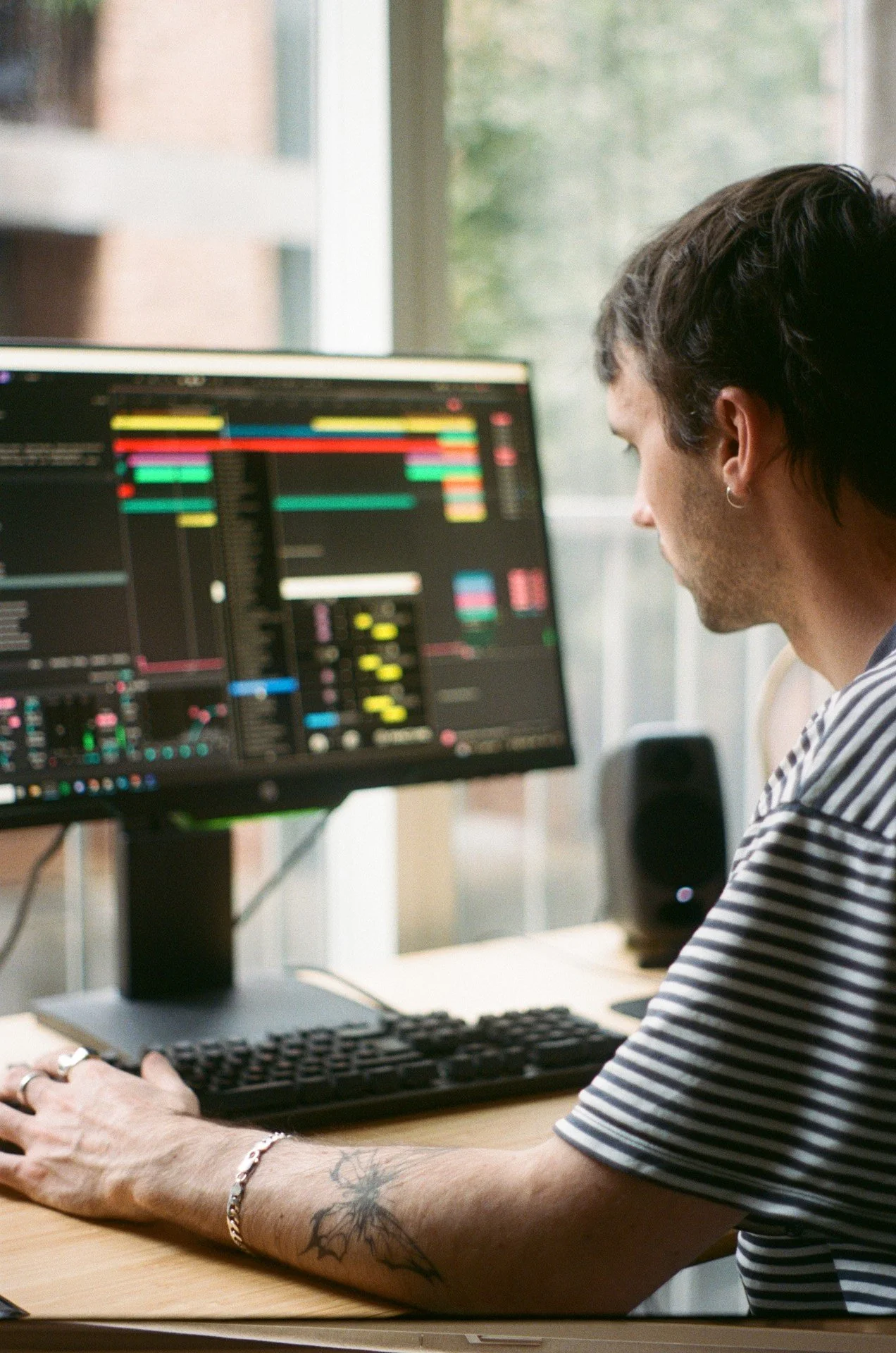Person working on computer with music production software open on screen, sitting at a desk near a window.