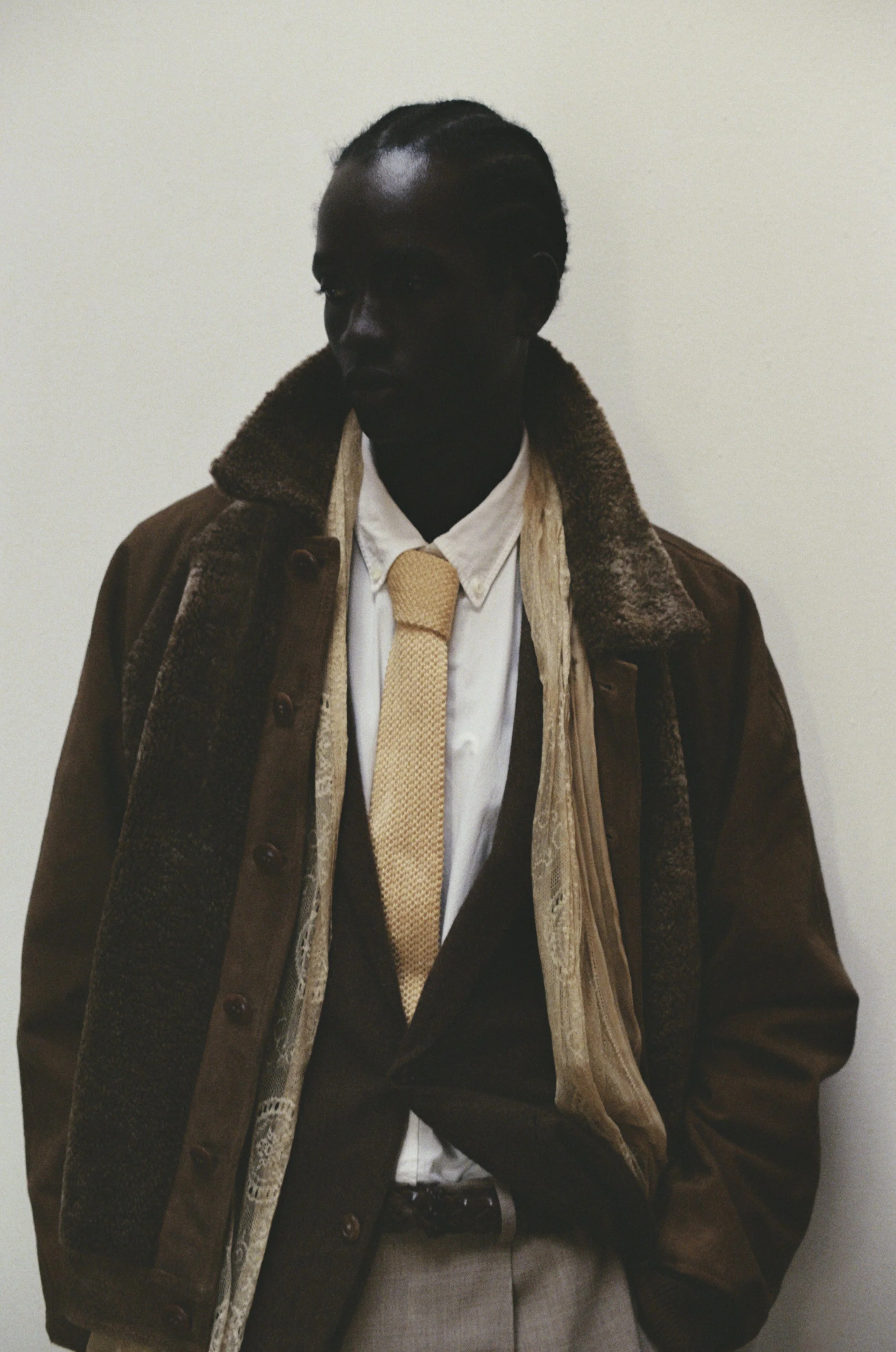 A person with dark skin and braided hair standing against a plain light-colored wall, dressed in vintage-style clothing including a brown coat with a shearling collar, a white shirt, a beige tie, and beige trousers.