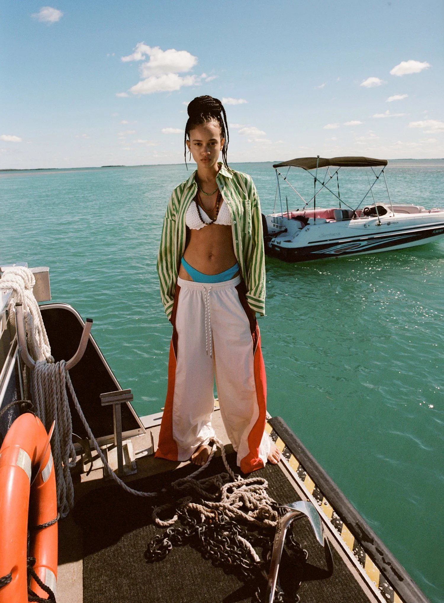 A woman with braided hair standing on a boat deck near the water, wearing a striped shirt, bikini top, and loose pants, with boats and a blue sky with clouds in the background.