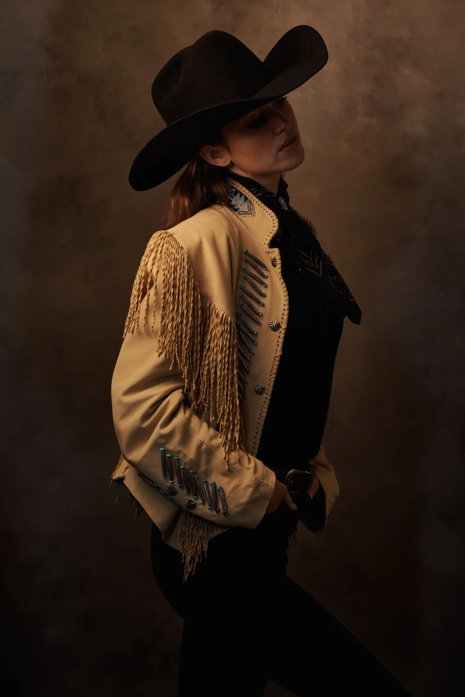 A woman wearing a large black cowboy hat, a beige fringe jacket with blue embellishments, and a black top, posed against a brown textured background.