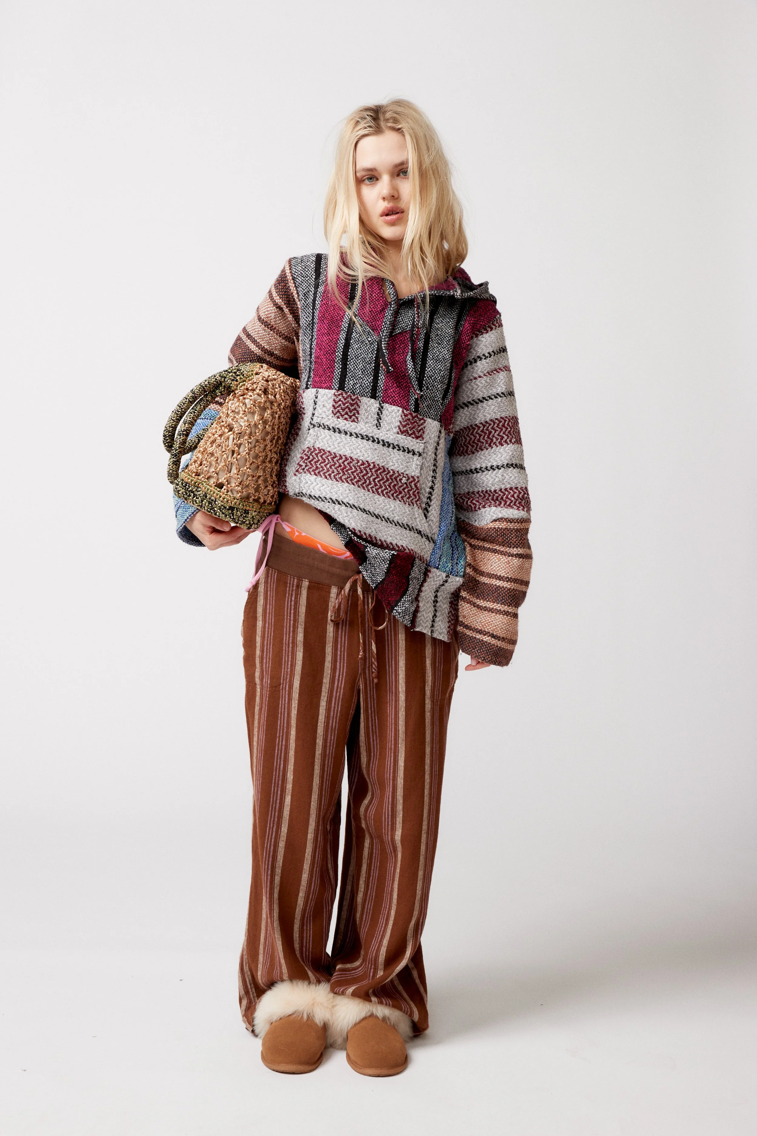 Young woman with blonde hair wearing a colorful, striped, quilted hoodie, striped brown and beige pants, and cozy fur-lined slippers, standing against a plain white background, holding a textured knit bag.