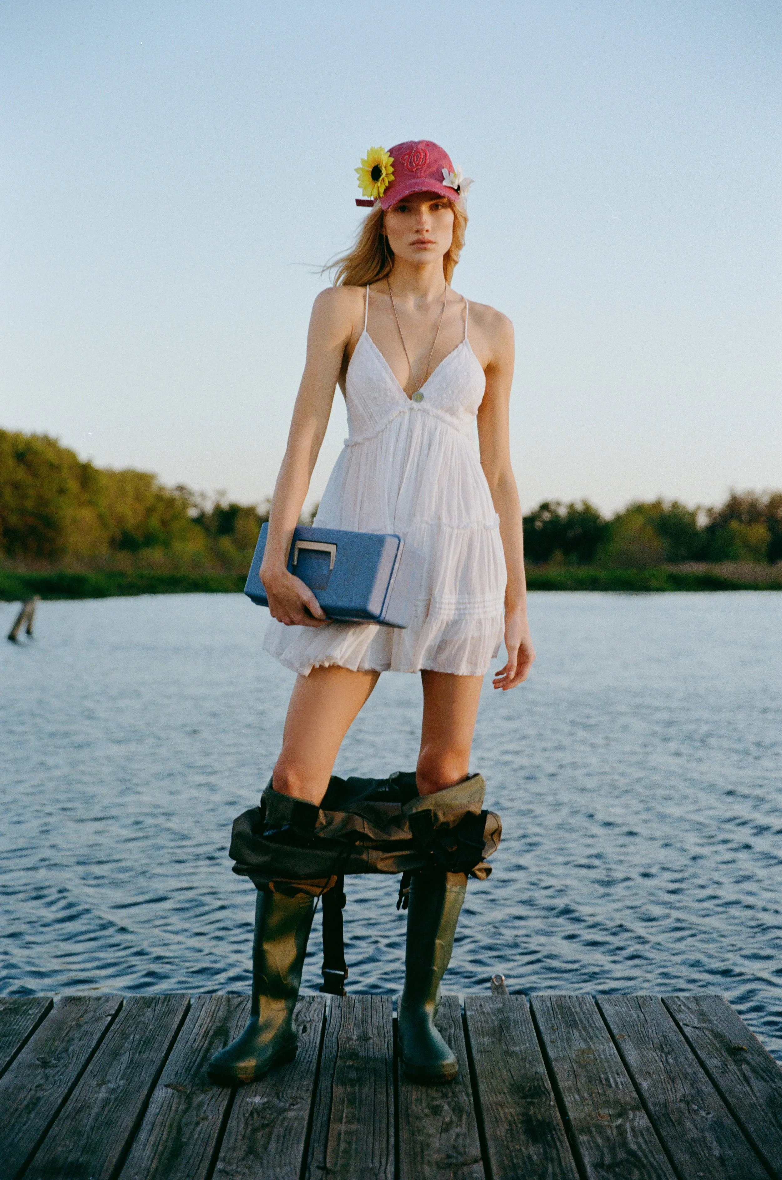 A young woman in a white dress with spaghetti straps stands on a wooden dock by the water, wearing a pink cap with a sunflower and Washington D.C. symbol, holding a blue cooler, and wearing camouflage rain boots.